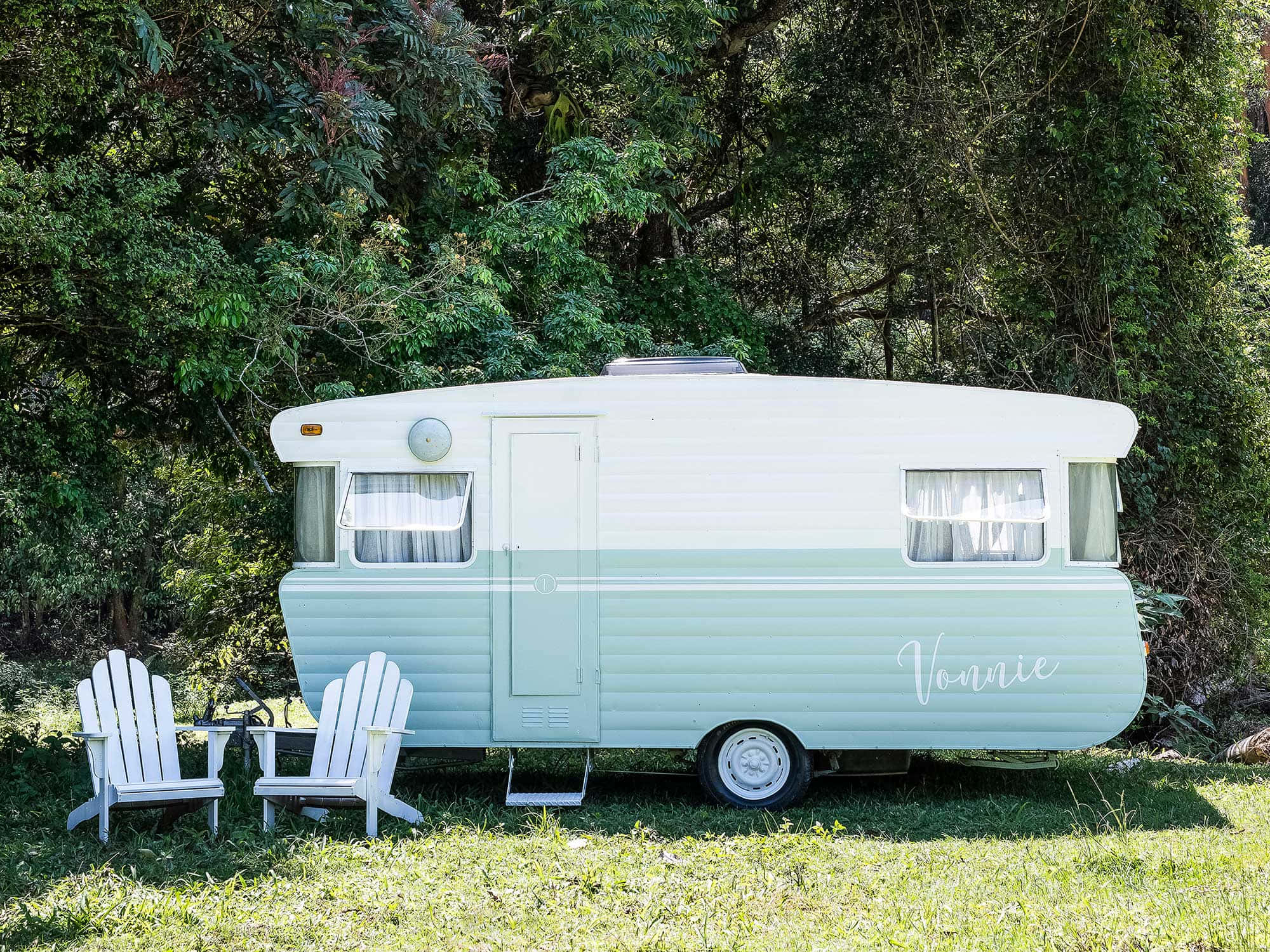 Caravan 桌布 壁紙 — 一輛名為「Vonnie」的復古薄荷綠和白色露營車，停放在陽光明媚的草地上，周圍是茂密的綠樹，附近有兩張白色阿迪朗達克椅子。