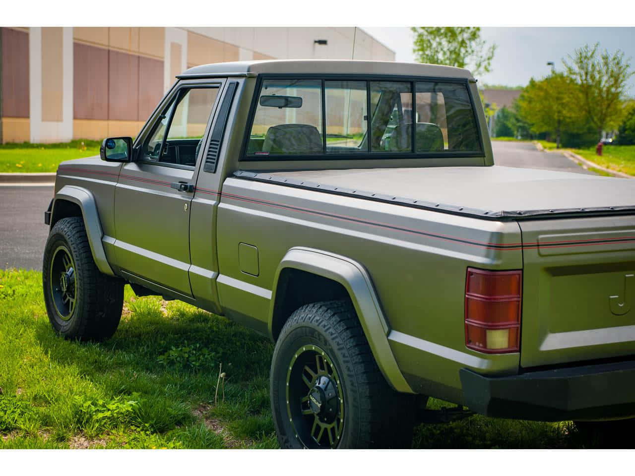 Jeep Comanche 桌布 壁紙 — 一輛啞光橄欖綠經典皮卡車，配黑色輪圈，在明亮的日光下停在路邊的草地上。