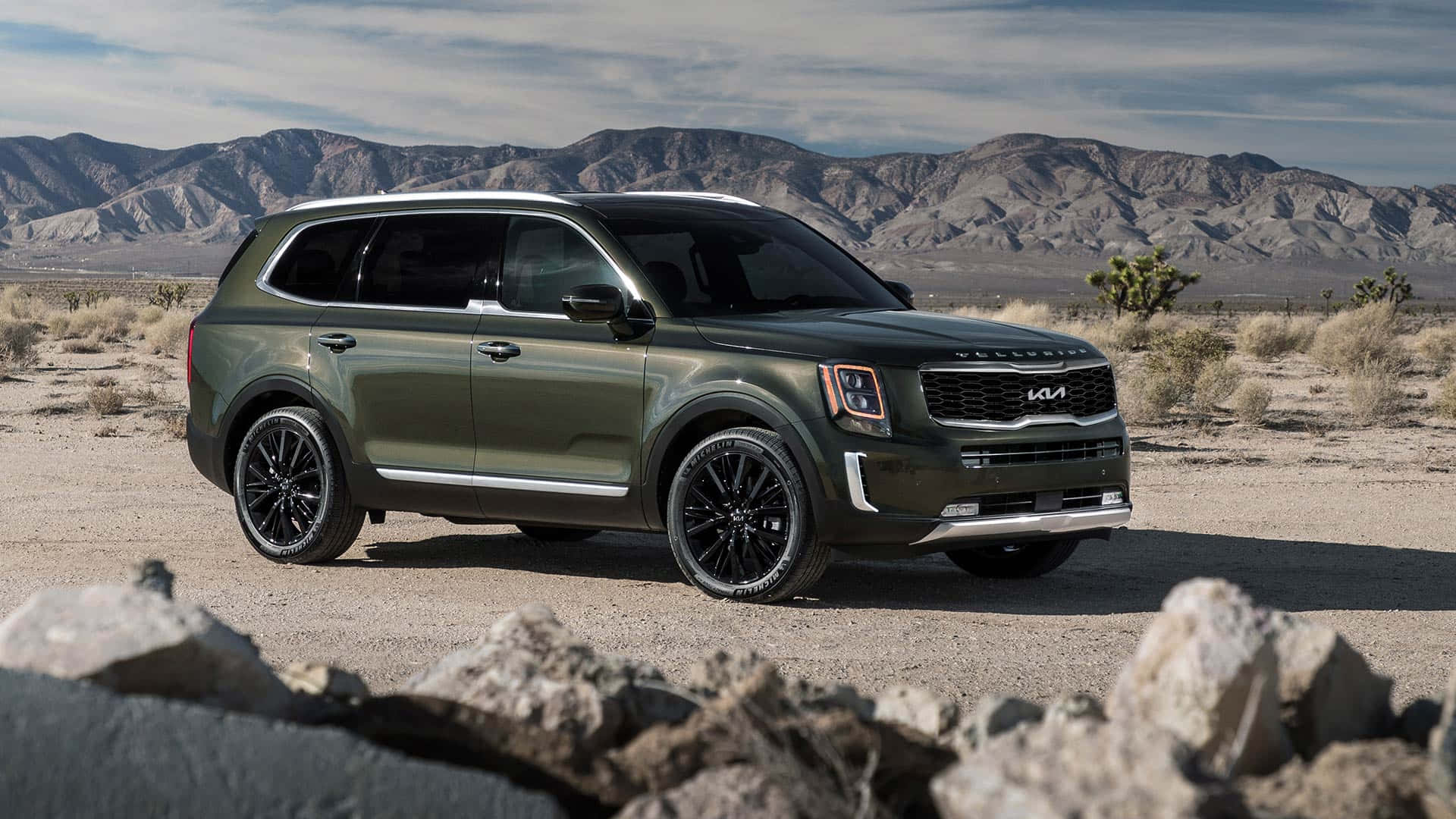 Kia Telluride 桌布 壁紙 — 一輛啞光橄欖綠色的Kia SUV停泊在岩石沙漠道路上，背景是崎嶇的山脈，天空部分多雲。