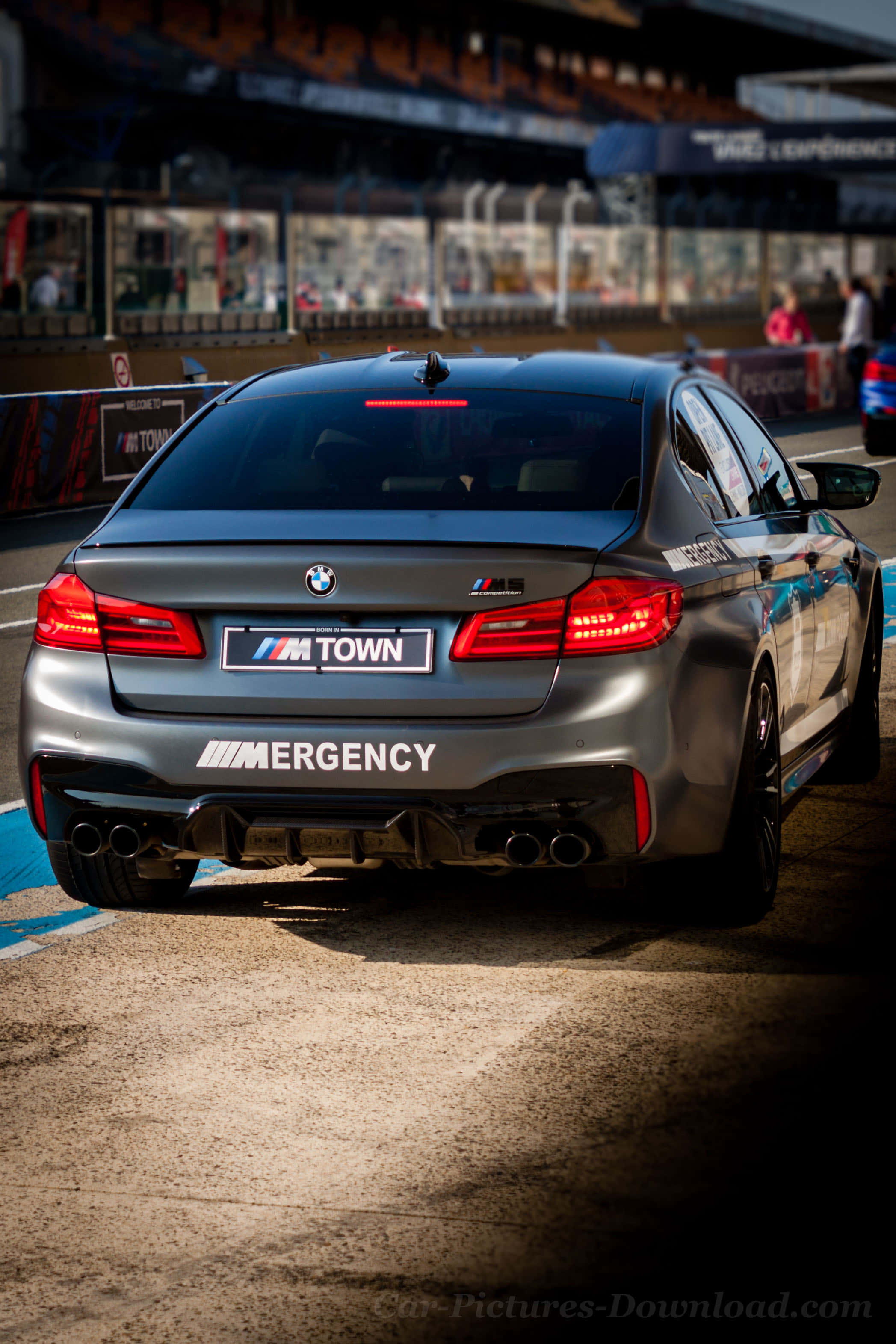 桌布 壁紙 — 一輛啞光灰色BMW M5賽車帶有「M TOWN」和「MERGENCY」貼紙，停在賽道上，紅色尾燈亮起。