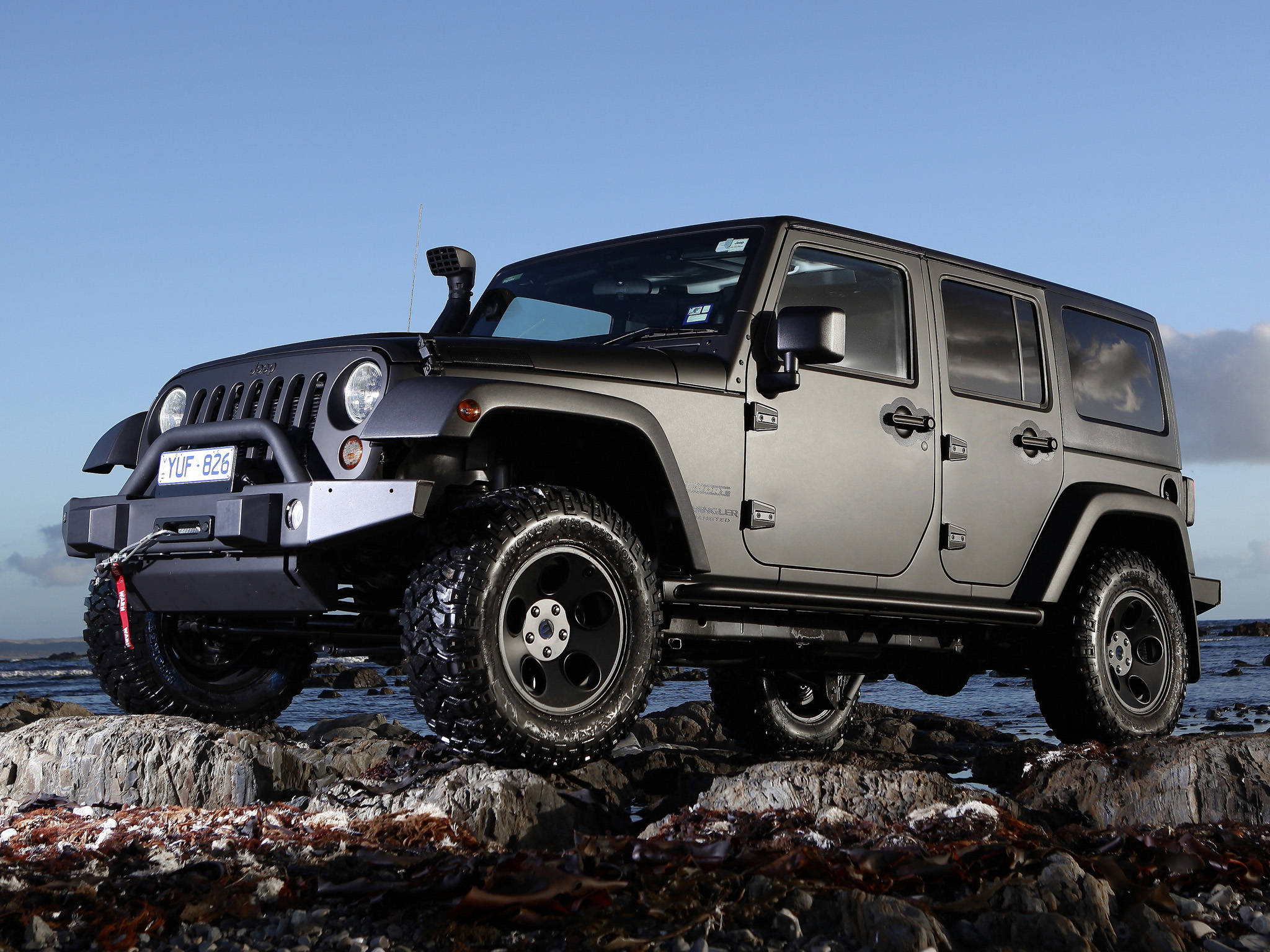 Black Jeep 桌布 壁紙 — 一輛啞光灰色Jeep Wrangler配黑色輪圈及前保險桿，在晴朗藍天下堅固地矗立於岩石海岸地形上。
