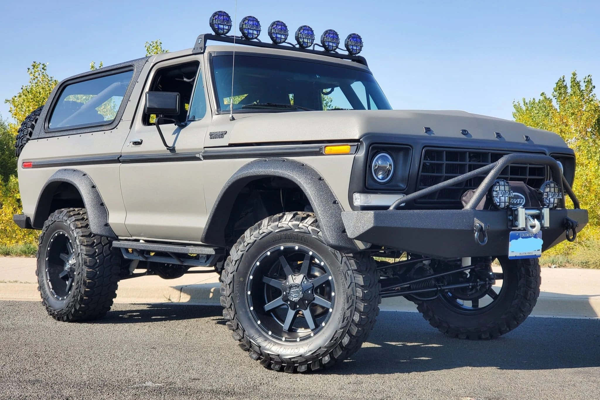 福特野馬越野車 桌布 壁紙 — 一輛啞光灰色升高的福特Bronco，配備超大輪胎和車頂探照燈，停在瀝青路面上，頭頂是晴朗的藍天。