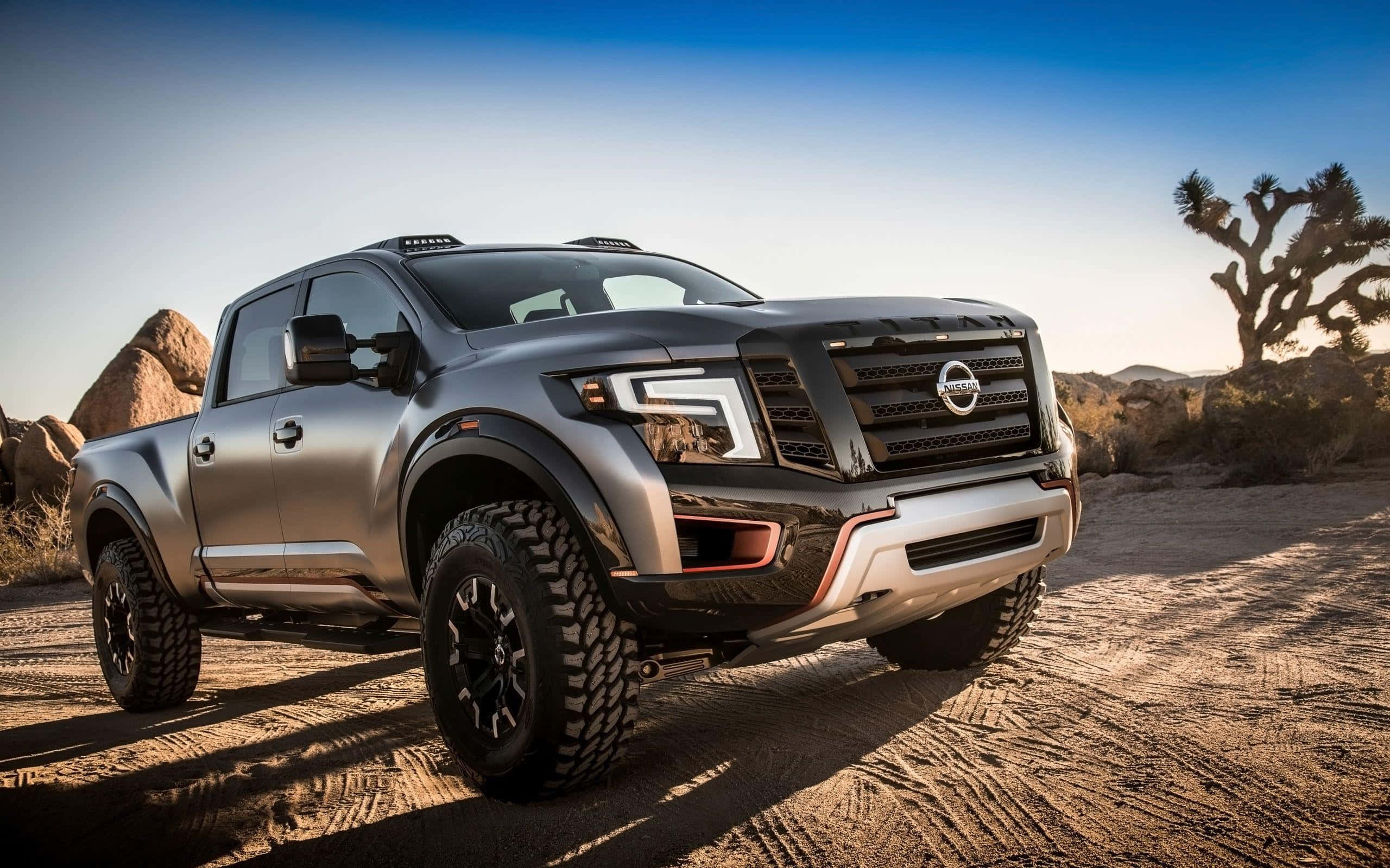 Nissan Titan 桌布 壁紙 — 一輛啞光灰色日產Frontier卡車，帶有黑色飾邊和大輪胎，停在沙質沙漠地形上，背景是晴朗的藍天和一棵約書亞樹。