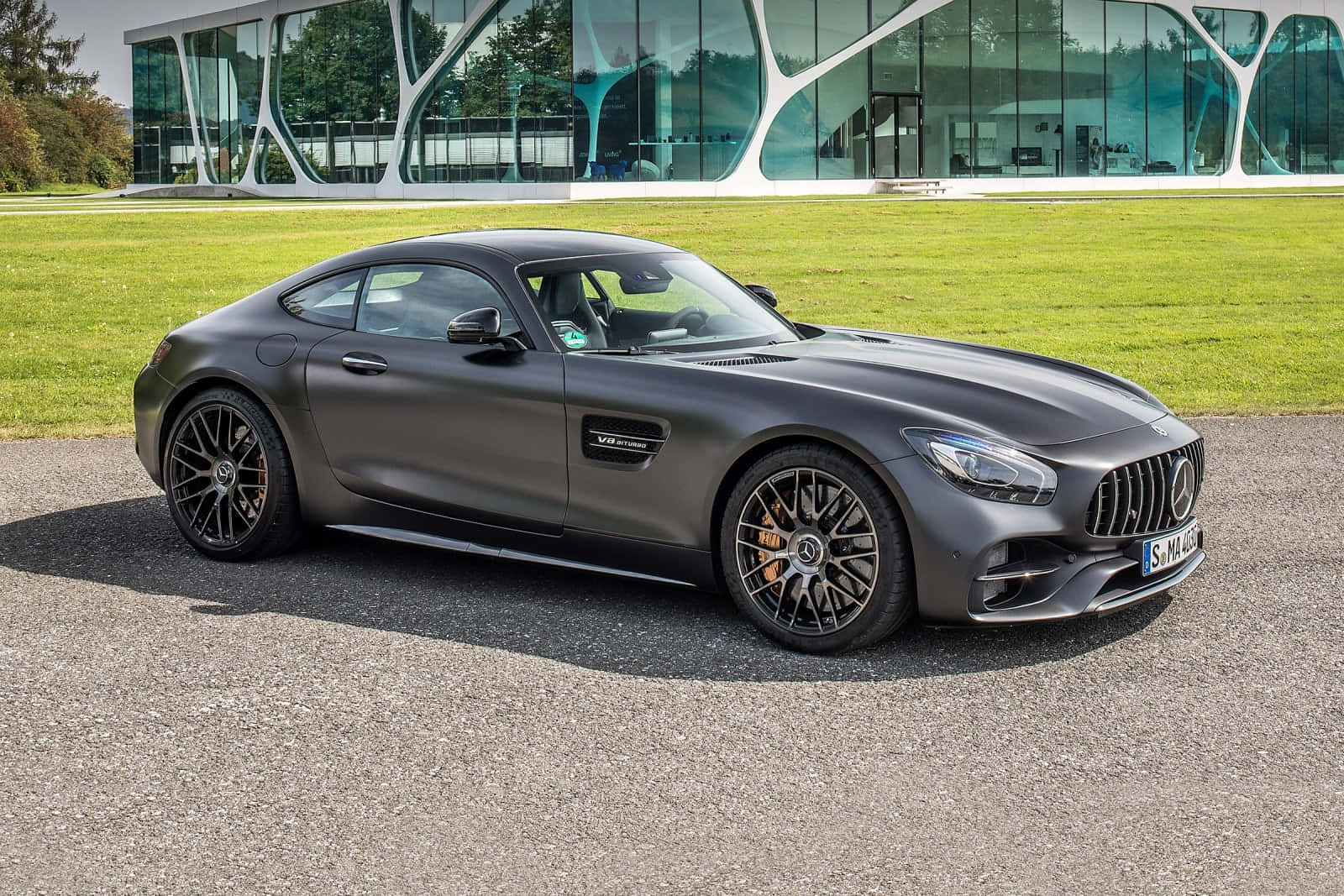 汽車 桌布 壁紙 — 一輛啞光灰色的 Mercedes-AMG GT 跑車停在柏油路上，前面是一棟有綠色草坪的現代玻璃建築。
