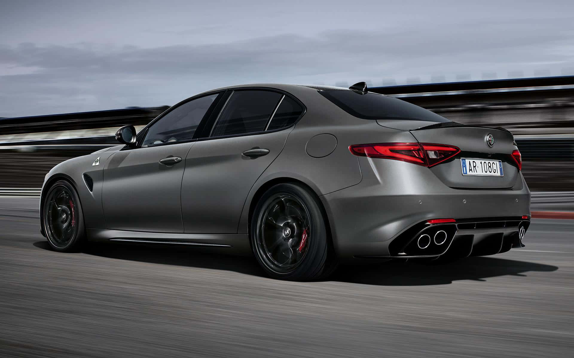 Alfa Romeo Giulia 桌布 壁紙 — 一輛啞光灰色的Alfa Romeo轎車在多雲的天空下，在模糊的賽道上疾馳。