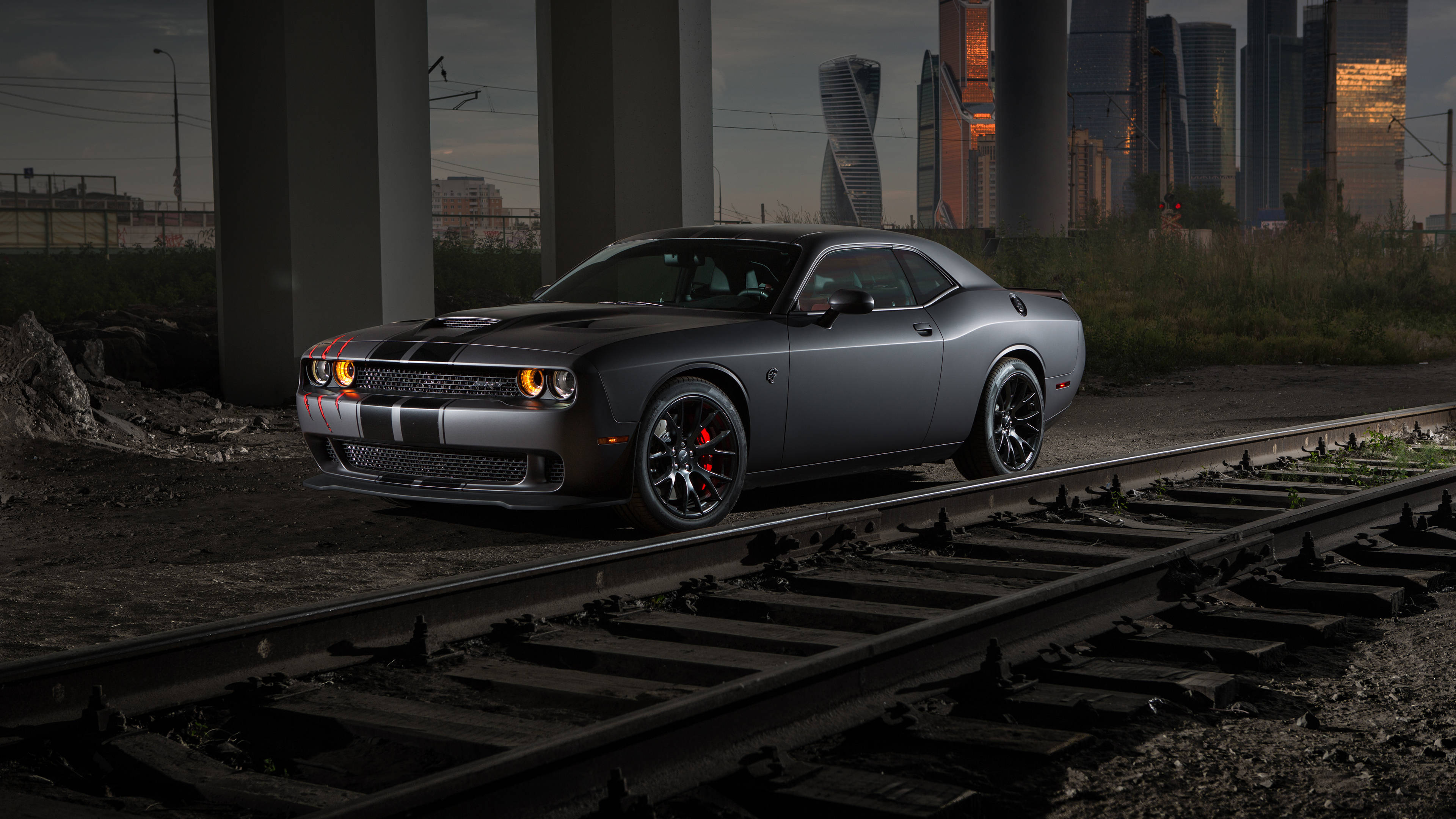 Dodge 桌布 壁紙 — 一輛啞光灰色的道奇挑戰者帶有紅色制動卡鉗，停放在生鏽的火車軌道上，位於高架橋下，城市天際線在黃昏時閃耀。