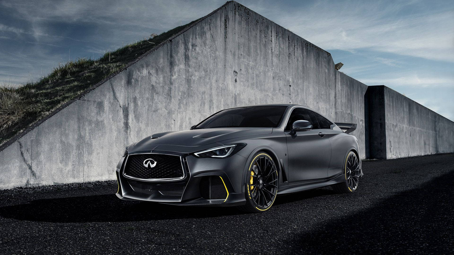 Infiniti 桌布 壁紙 — 一輛啞光灰色英菲尼迪跑車，帶有黃色點綴，停泊在陰天下一座高聳的混凝土牆旁。