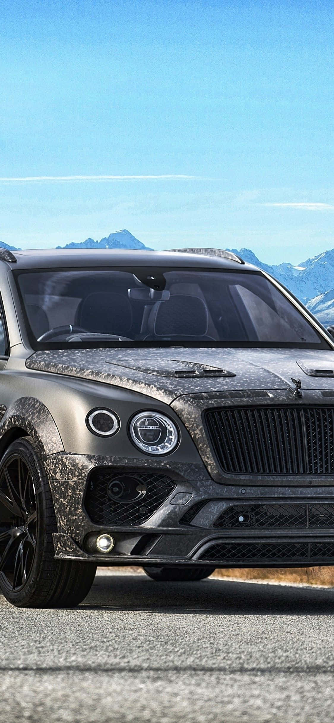 bentley HD 桌布 壁紙 — 一輛啞光灰色賓利Bentayga SUV，帶有迷彩包裹，停在山路上，天空清澈湛藍，遠處有白雪覆蓋的山峰。