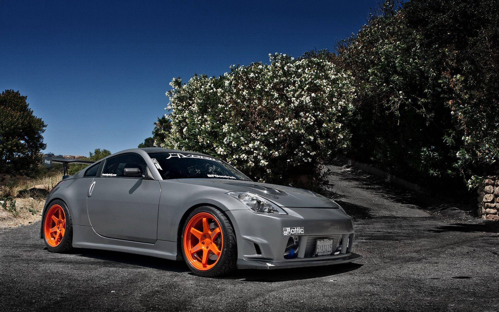 Nissan 350z 桌布 壁紙 — 一輛啞光灰色跑車配以鮮豔橙色車輪，停泊在碎石車道上，旁邊是盛開的白色灌木叢，上方是清澈的藍天。