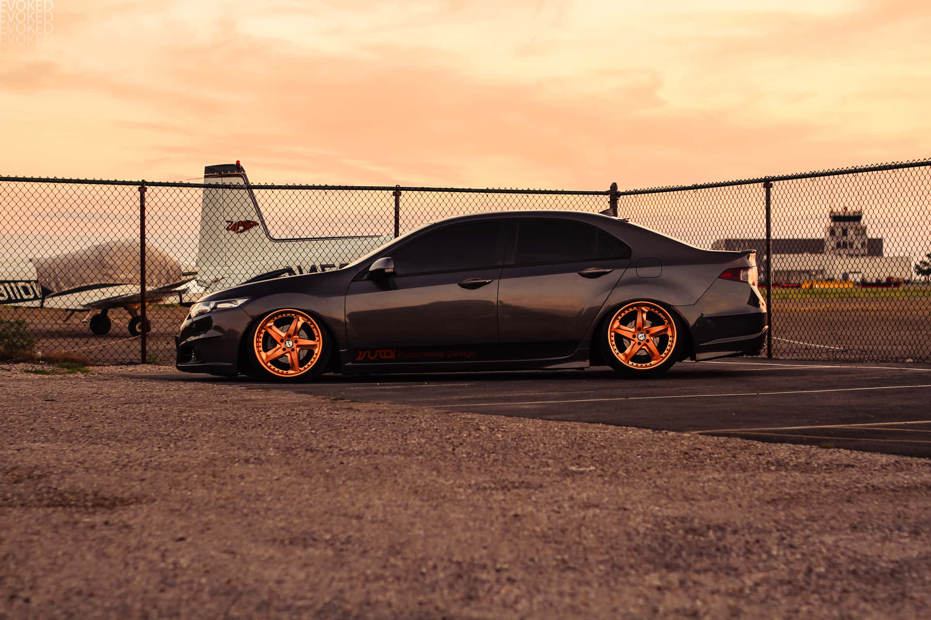 Acura Tsx 桌布 壁紙 — 一輛啞光灰色轎車，配備發光橙色輪圈，停泊在日落時分的碎石停車場，背景有鐵絲網圍欄和飛機。