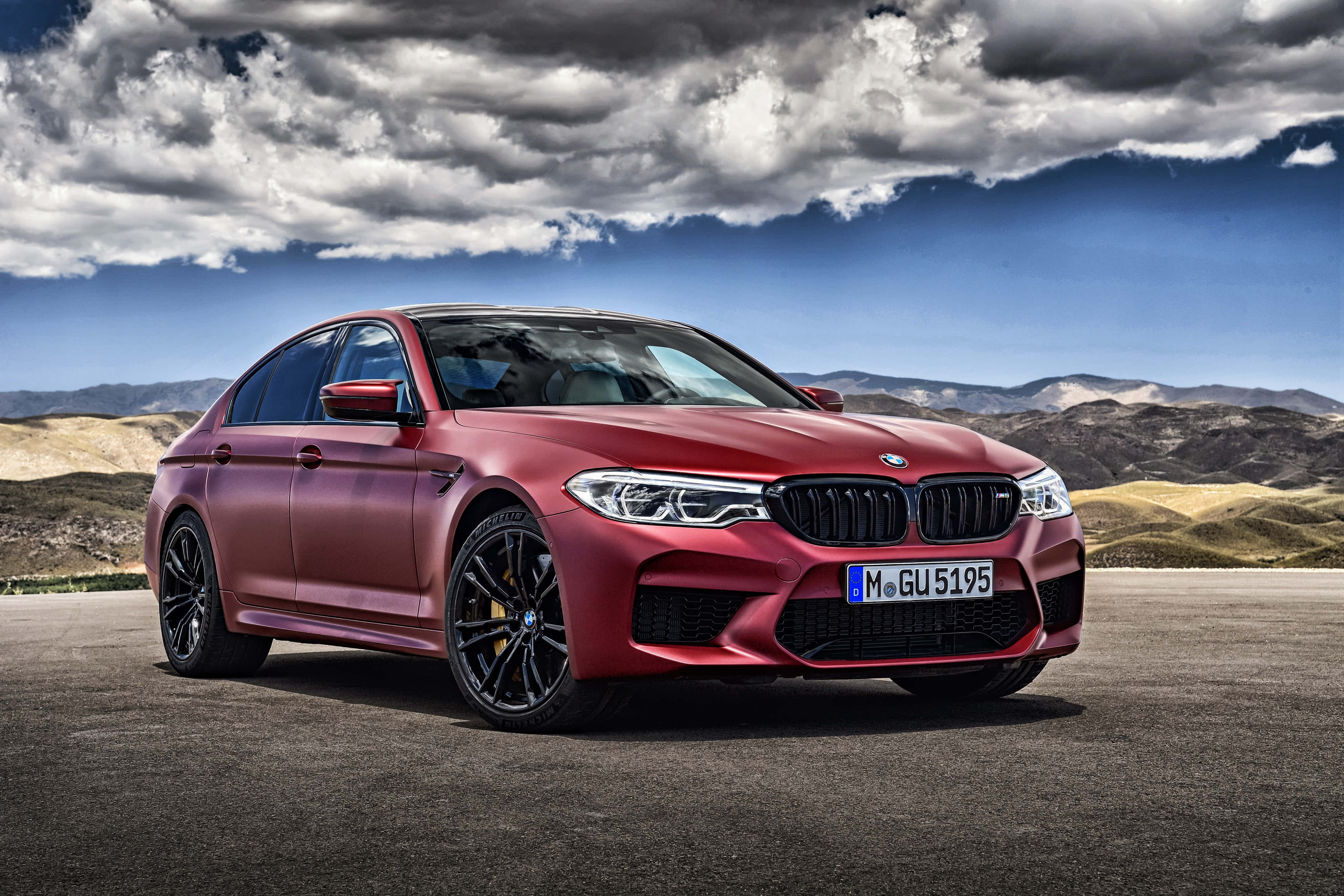 桌布 壁紙 — 一輛啞光紅色的BMW M5運動轎車停在瀝青路上，背景有戲劇性的雲層和山區地形。