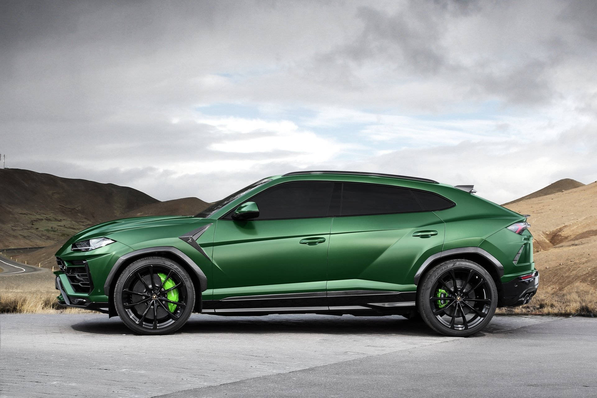 lambo 桌布 壁紙 — 一輛啞光綠色的藍寶堅尼 Urus SUV 停放在鋪砌道路上，背景是起伏的棕色山丘，天空多雲。