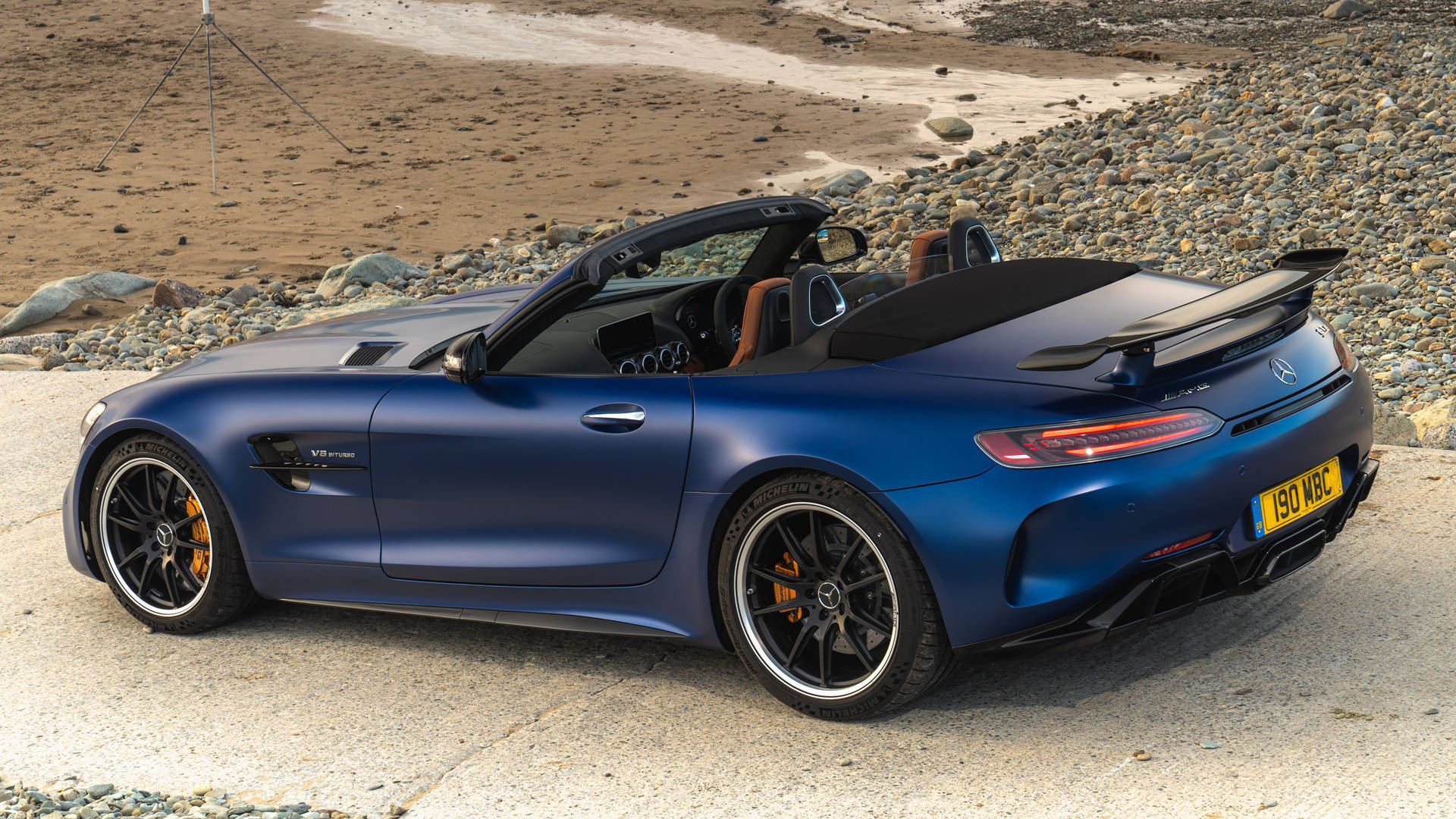 R 桌布 壁紙 — 一輛啞光藍色Mercedes-AMG GT Roadster停放在岩石海灘旁的混凝土板上，背景有一個三腳架。