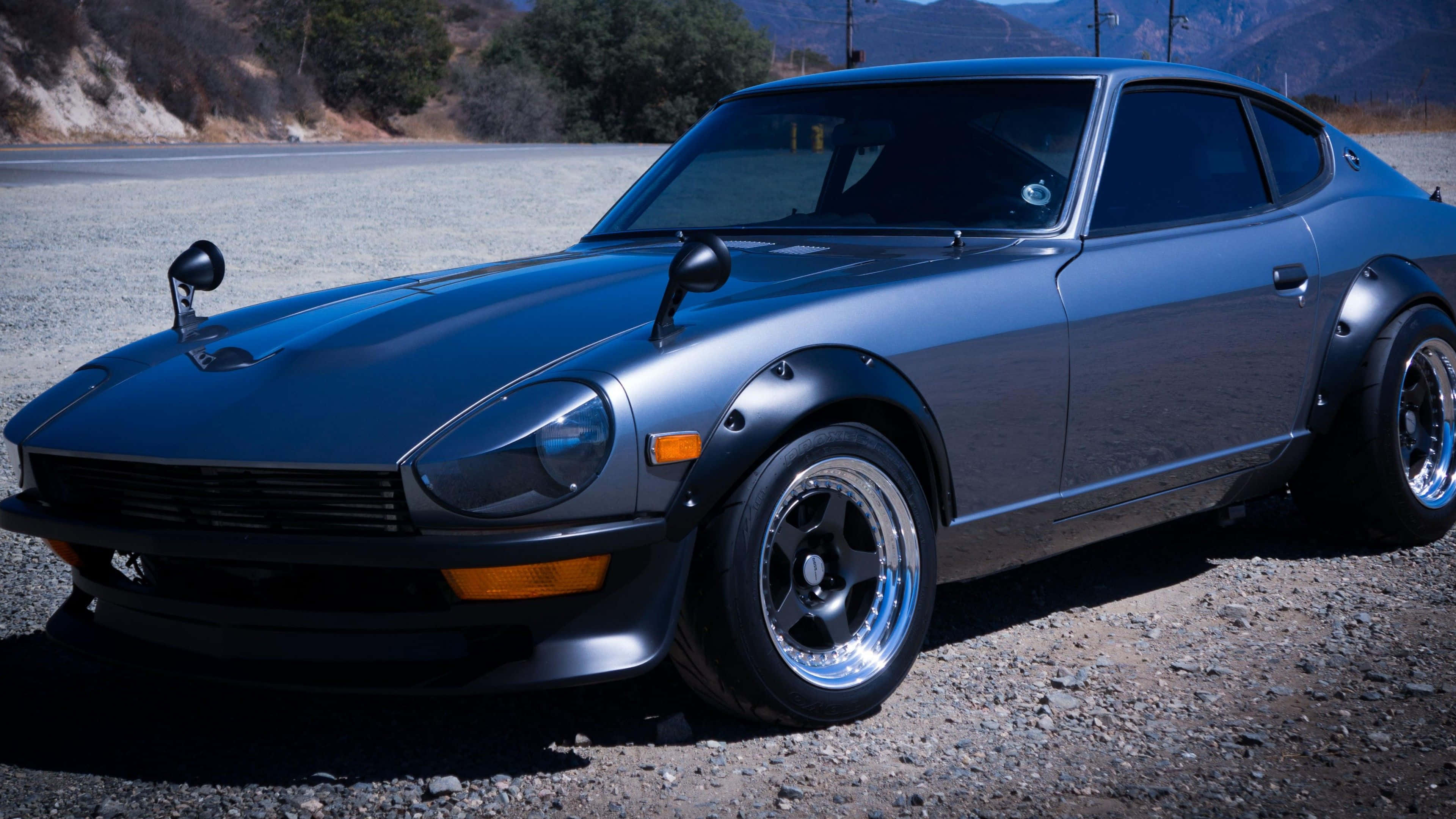 Datsun 桌布 壁紙 — 一輛啞光藍色經典跑車配備定制輪圈，在明亮日光下停泊在沙漠道路旁的礫石上。