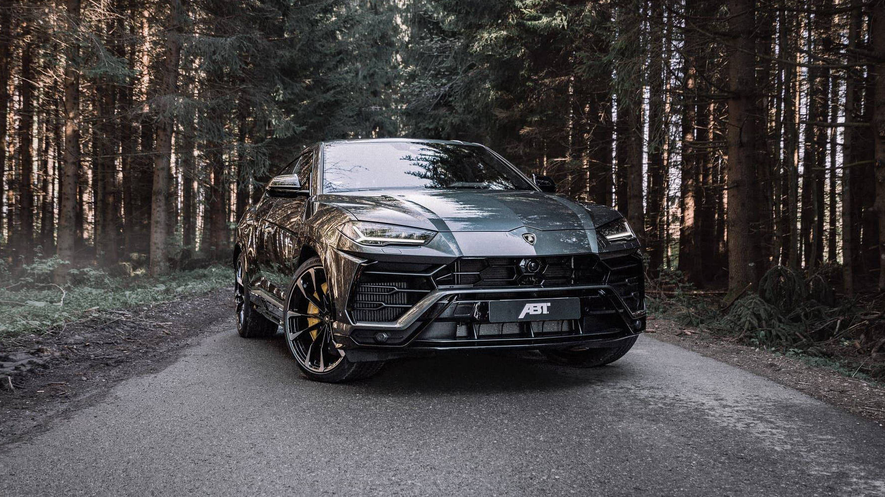 美學 桌布 壁紙 — 一輛啞光黑色ABT藍寶堅尼Urus SUV，帶金色點綴，停泊在松樹環繞的森林道路上。