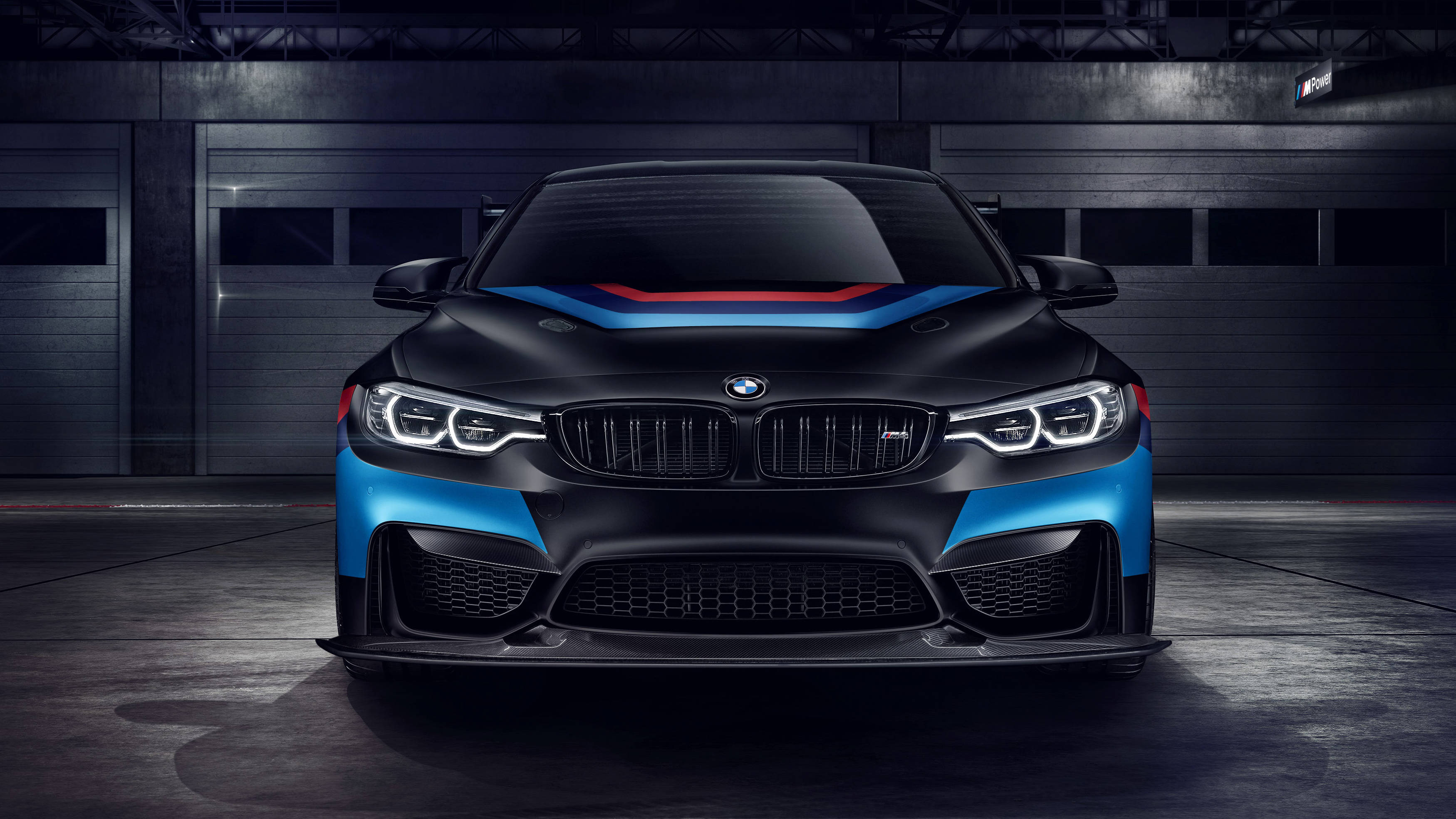 m 桌布 壁紙 — 一輛啞光黑色BMW M2帶有醒目的藍色和紅色賽車條紋，在昏暗的工業車庫中直視前方。