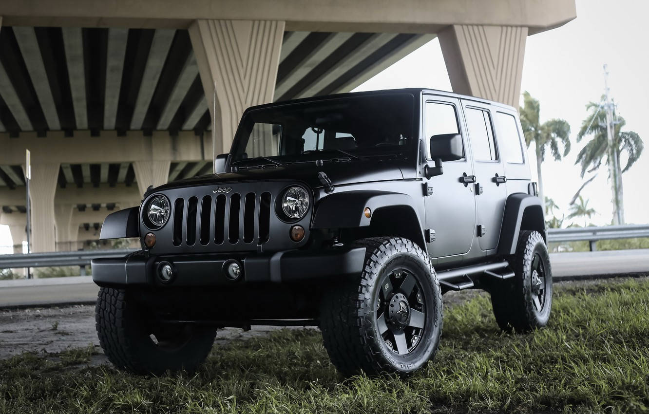 Black Jeep 桌布 壁紙 — 一輛啞光黑色Jeep Wrangler停在混凝土天橋下的草地上，背景有棕櫚樹。