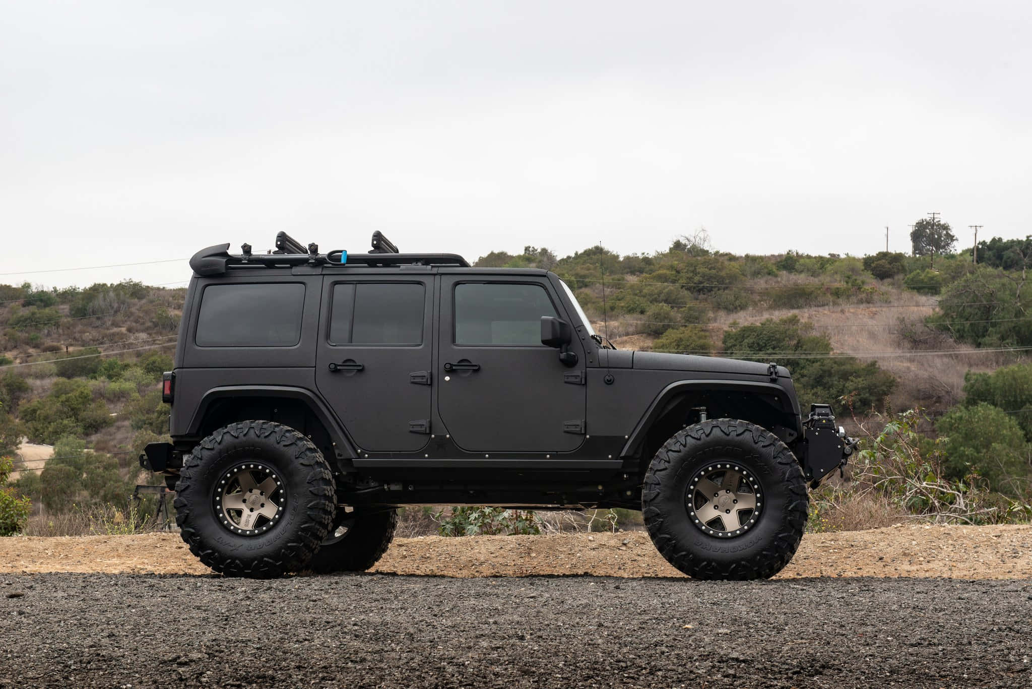 黑色吉普車 桌布 壁紙 — 一輛啞光黑色升高的Jeep Wrangler，配備超大輪胎，停放在碎石上，旁邊是灌木覆蓋的小山丘，天空多雲。