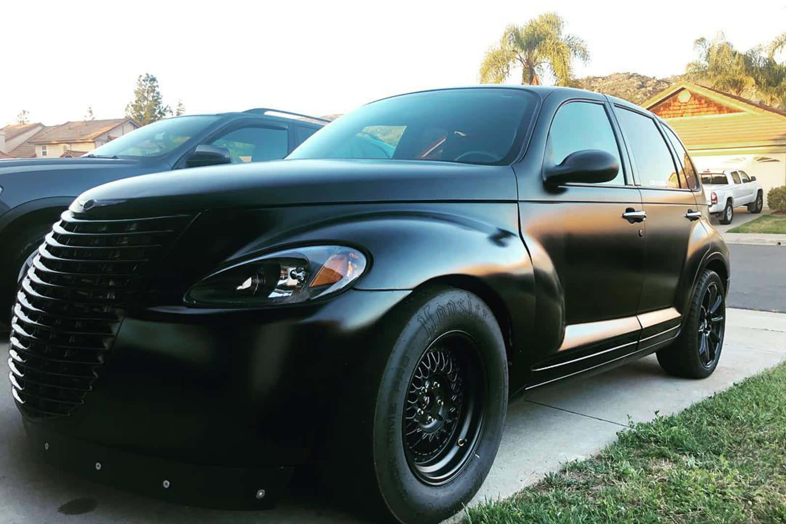 Chrysler Pt Cruiser 桌布 壁紙 — 一輛啞光黑色的克萊斯勒 PT Cruiser 配備定制輪圈，在黃金時刻停放在郊區的車道上。