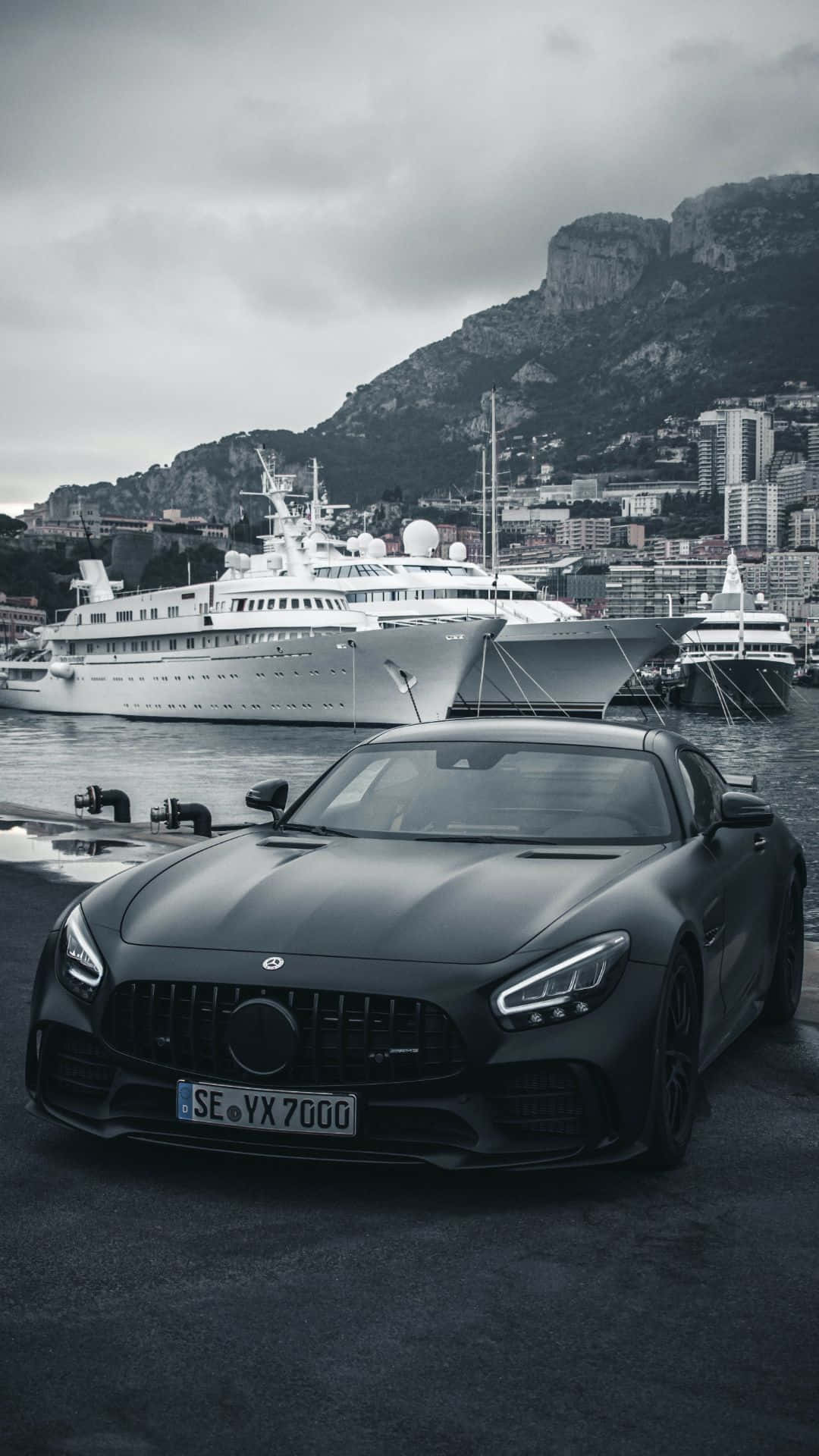跑車 桌布 壁紙 — 一輛啞光黑色的梅賽德斯-AMG GT停泊在碼頭，背景有豪華遊艇和懸崖城市，天空多雲。