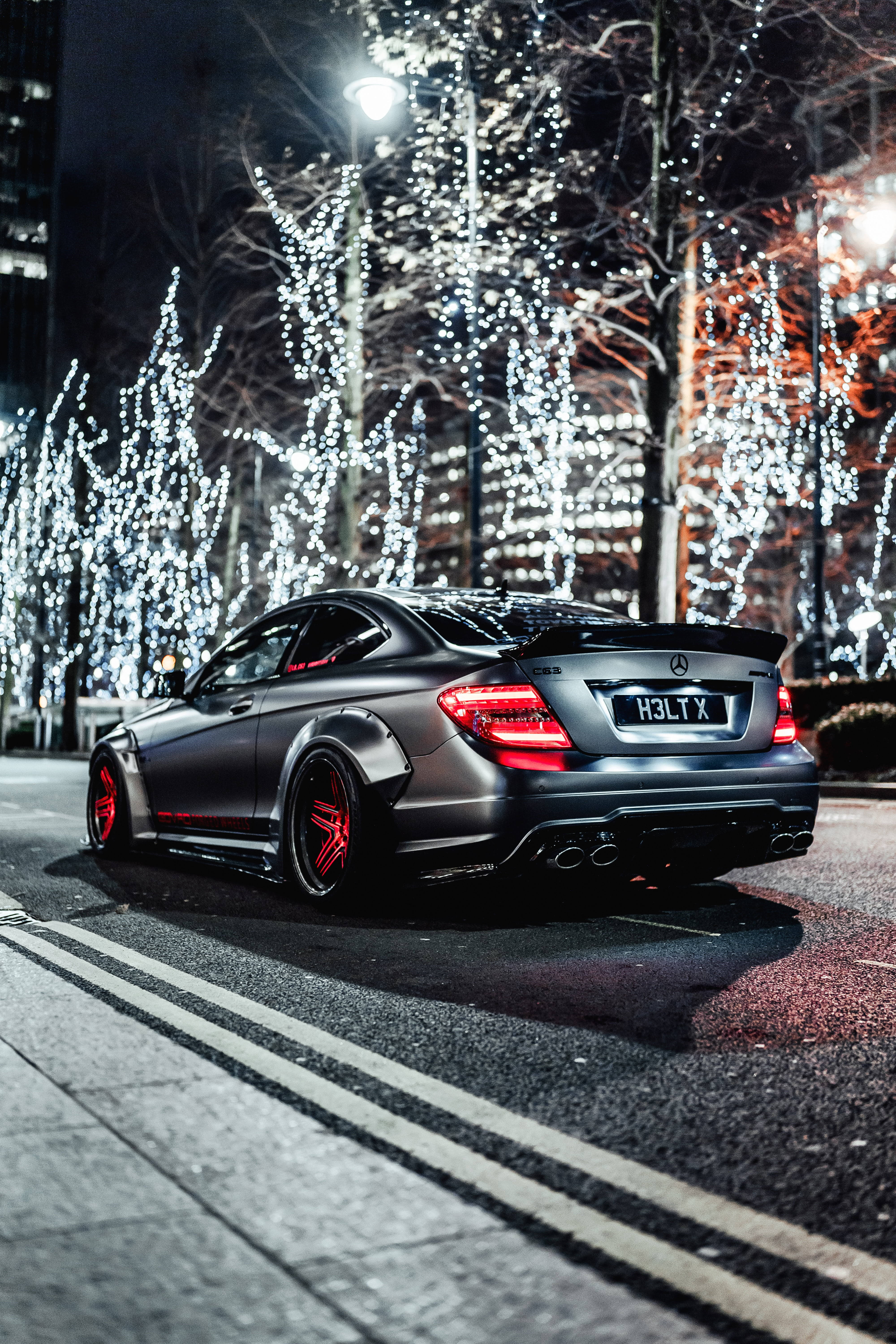 mercedes amg iPhone 桌布 壁紙 — 一輛啞光黑色的賓士雙門轎跑車帶有紅色點綴，夜晚停泊在城市的街道上，光禿禿的樹上掛著發光的燈串。