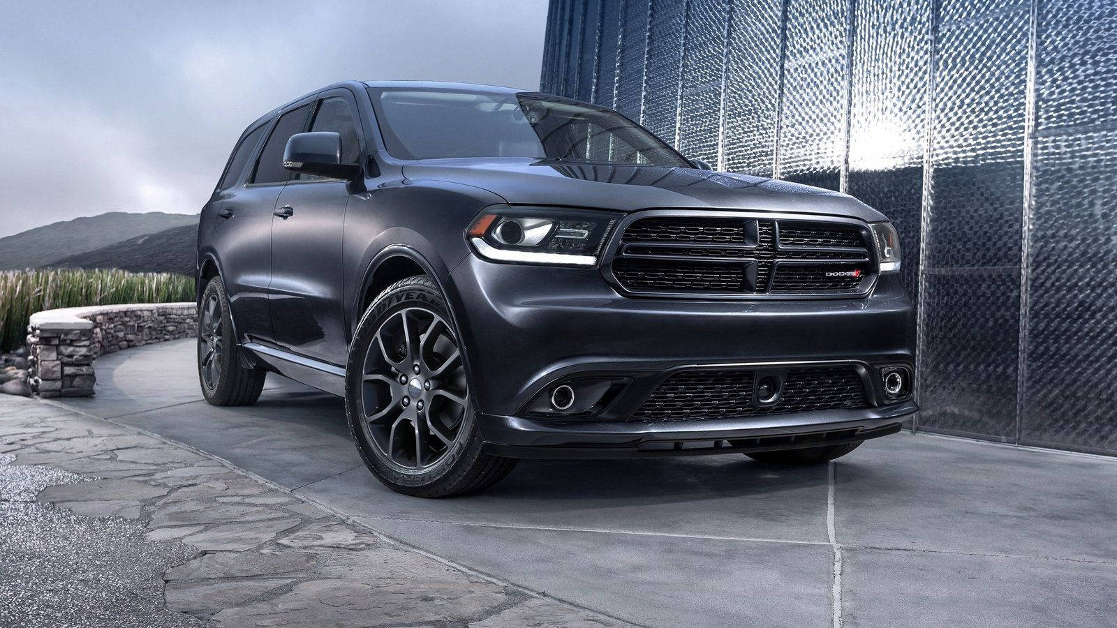 Dodge 桌布 壁紙 — 一輛啞光黑色的道奇杜蘭戈SUV停放在石頭車道上，旁邊是帶紋理的金屬牆，背景有山丘。