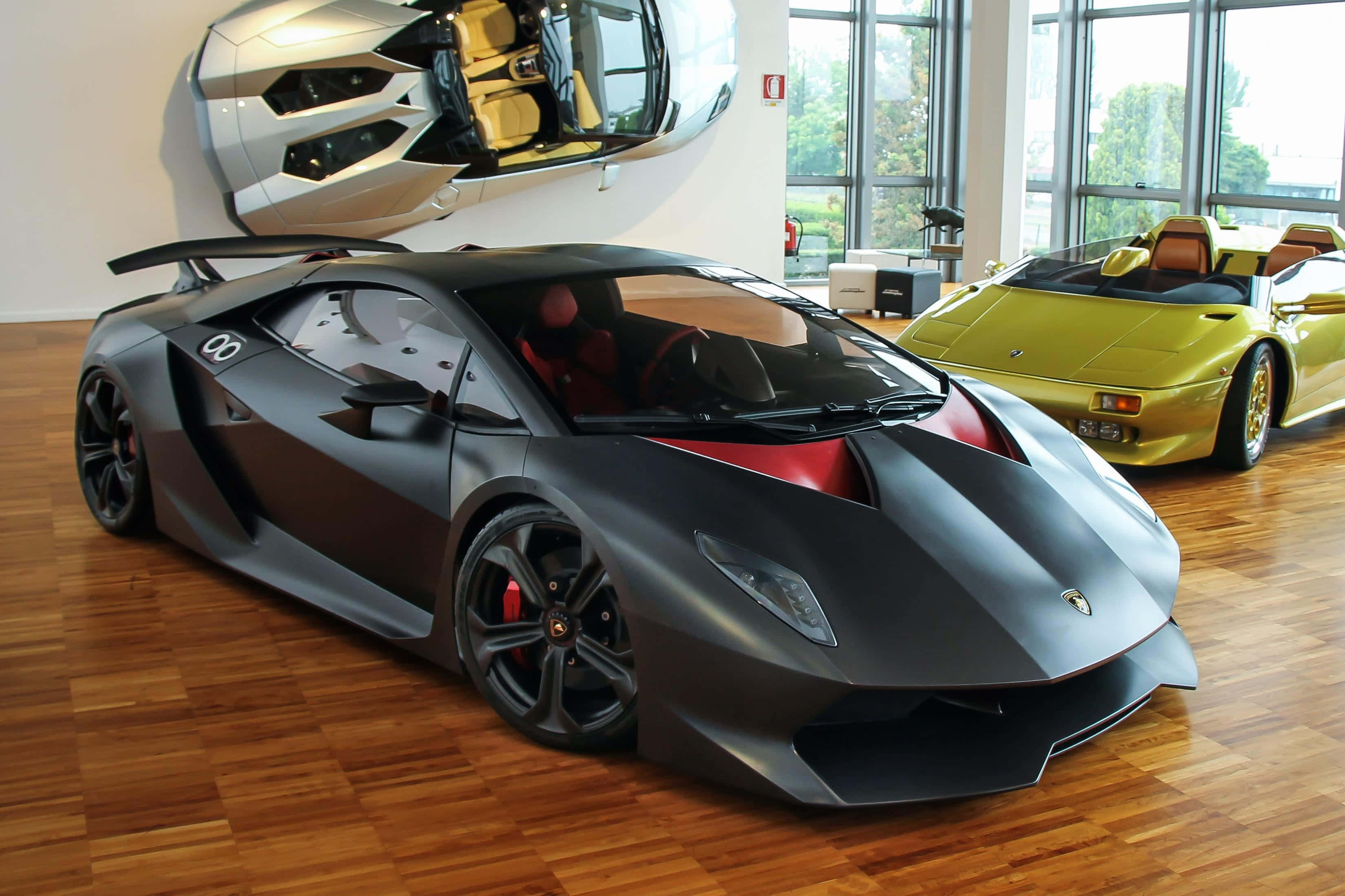 Lamborghini Sesto Elemento 桌布 壁紙 — 一輛啞光黑色藍寶堅尼跑車帶有紅色點綴，突出地放置在明亮展廳的木地板上，背景有一輛黃色敞篷車和一個壁掛式汽車雕塑。