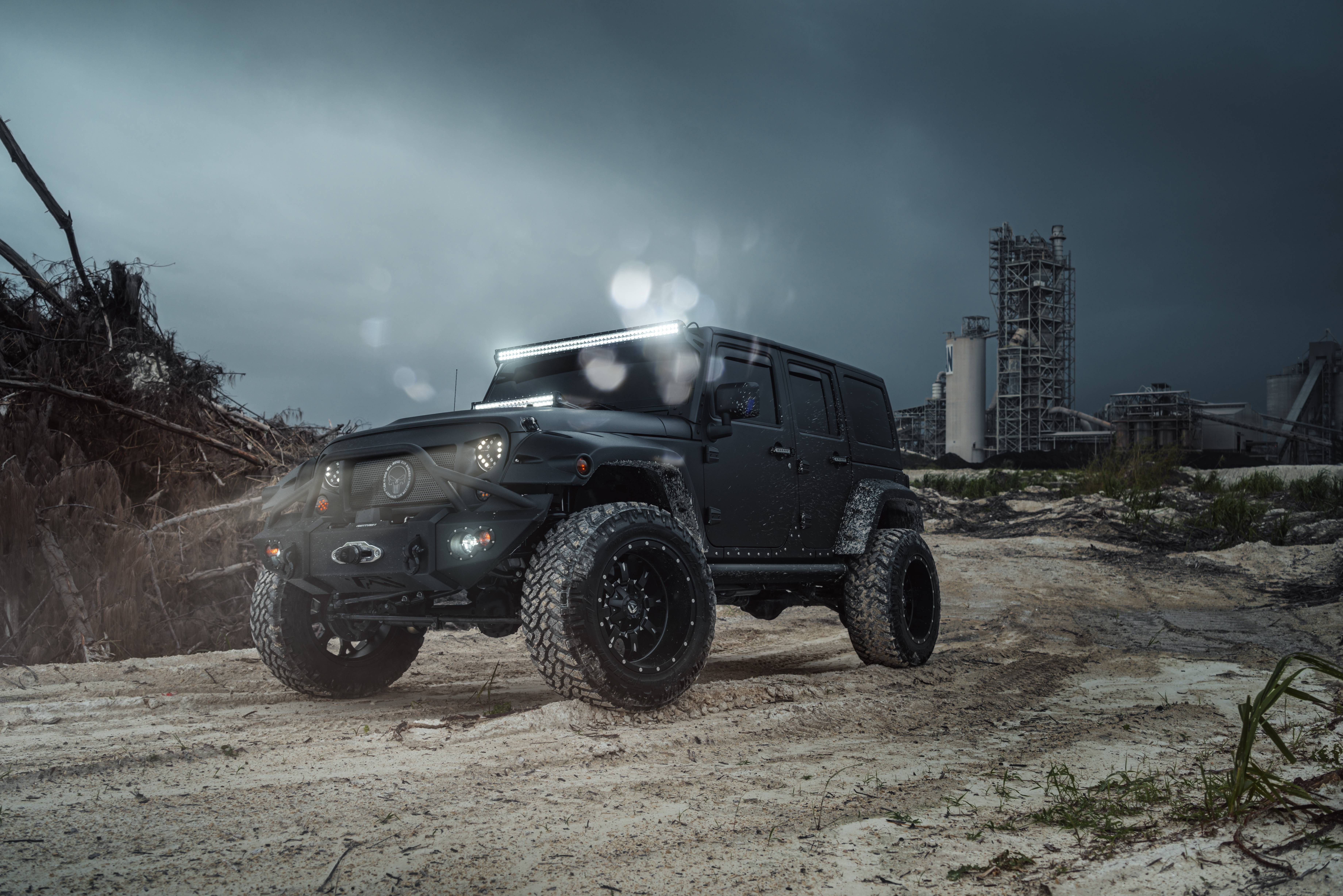 Wrangler 桌布 壁紙 — 一輛啞光黑色越野Jeep配備超大輪胎，在暴風雨的天空下駛過荒涼的工業廢墟。