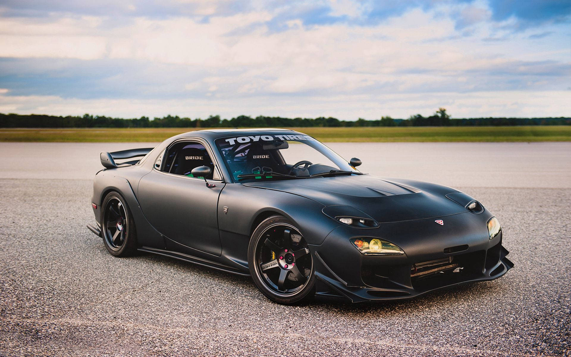 Mazda 桌布 壁紙 — 一輛啞光黑色馬自達RX-7跑車，配備Toyo輪胎和Bride座椅，停在碎石路上，天空多雲。