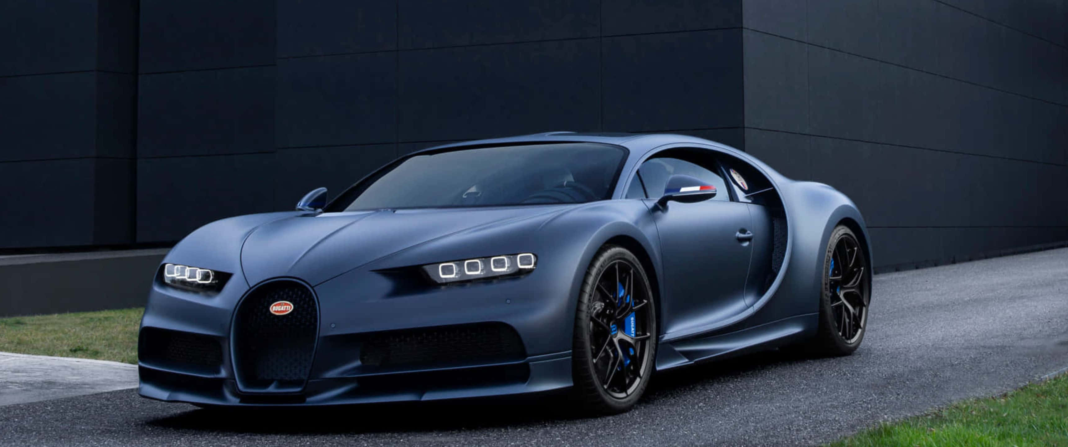3440x1440p Bugatti 桌布 壁紙 — 一輛啞藍色Bugatti Chiron跑車停在柏油路上，旁邊是有草叢的深色牆壁。