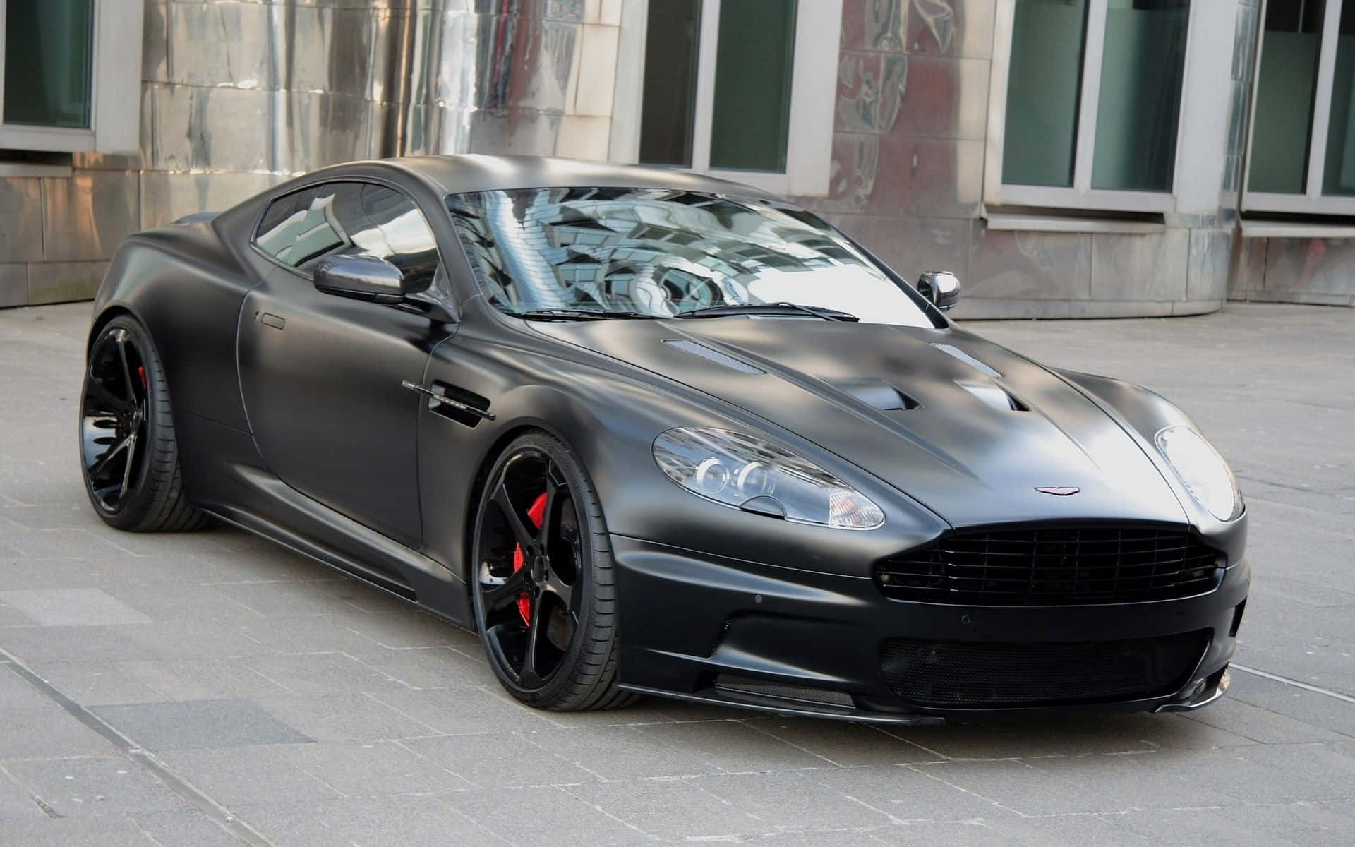 Aston Martin Db9 桌布 壁紙 — 一輛啞黑色 Aston Martin 跑車配備紅色煞車卡鉗，停放在磁磚鋪設的城市人行道上。