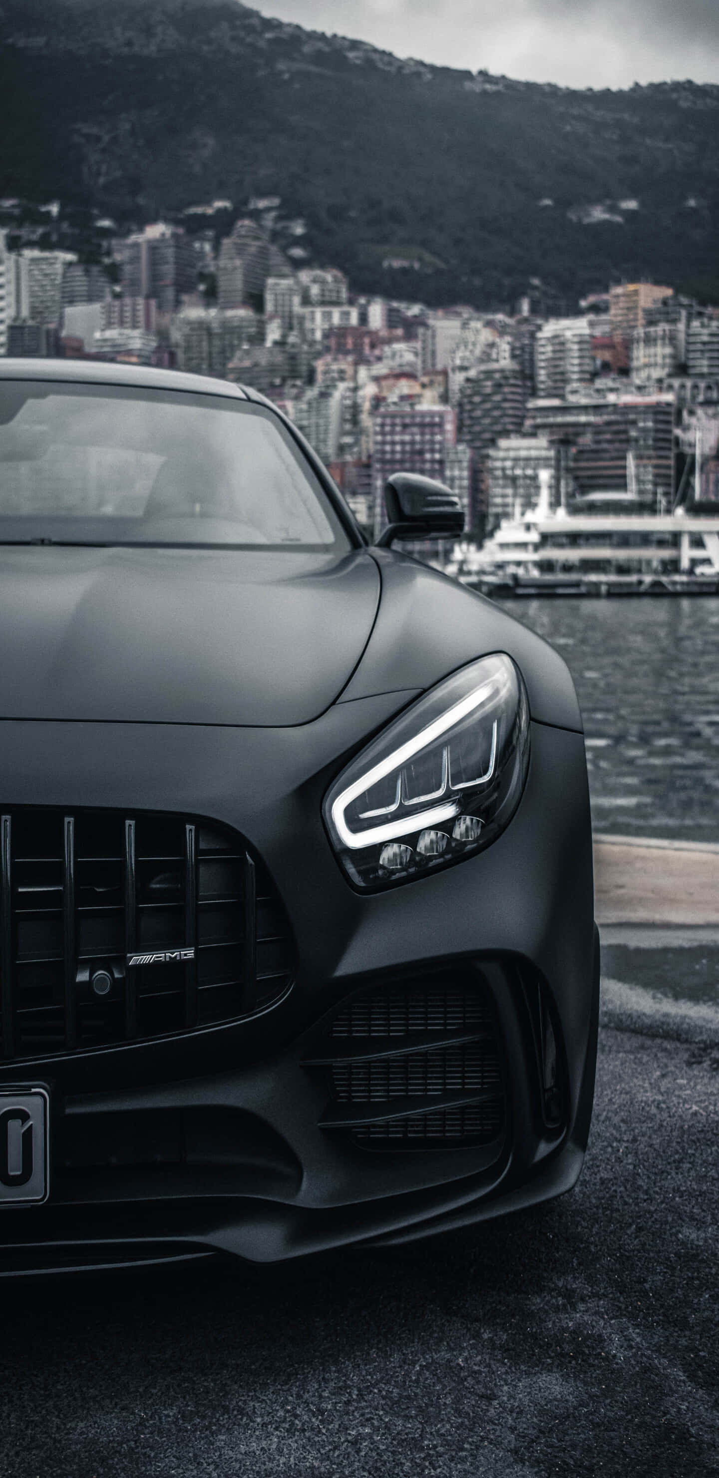 Car Images HD 桌布 壁紙 — 一輛啞黑色AMG跑車停泊在海濱，背景有城市景觀和山脈。