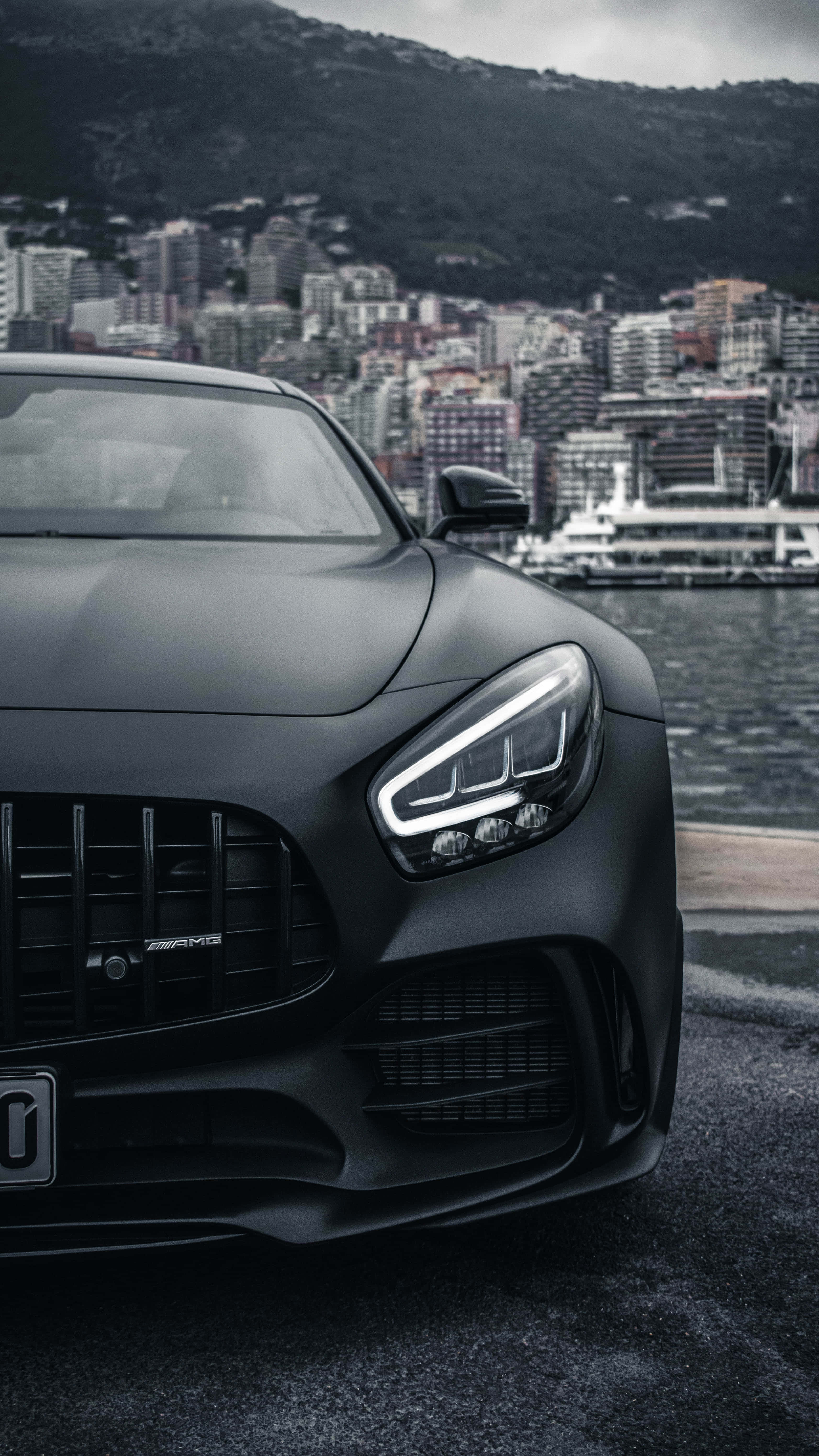 Mercedes 桌布 壁紙 — 一輛啞黑色AMG跑車停泊在海濱，背景是城市景觀和山脈，天色陰沉。