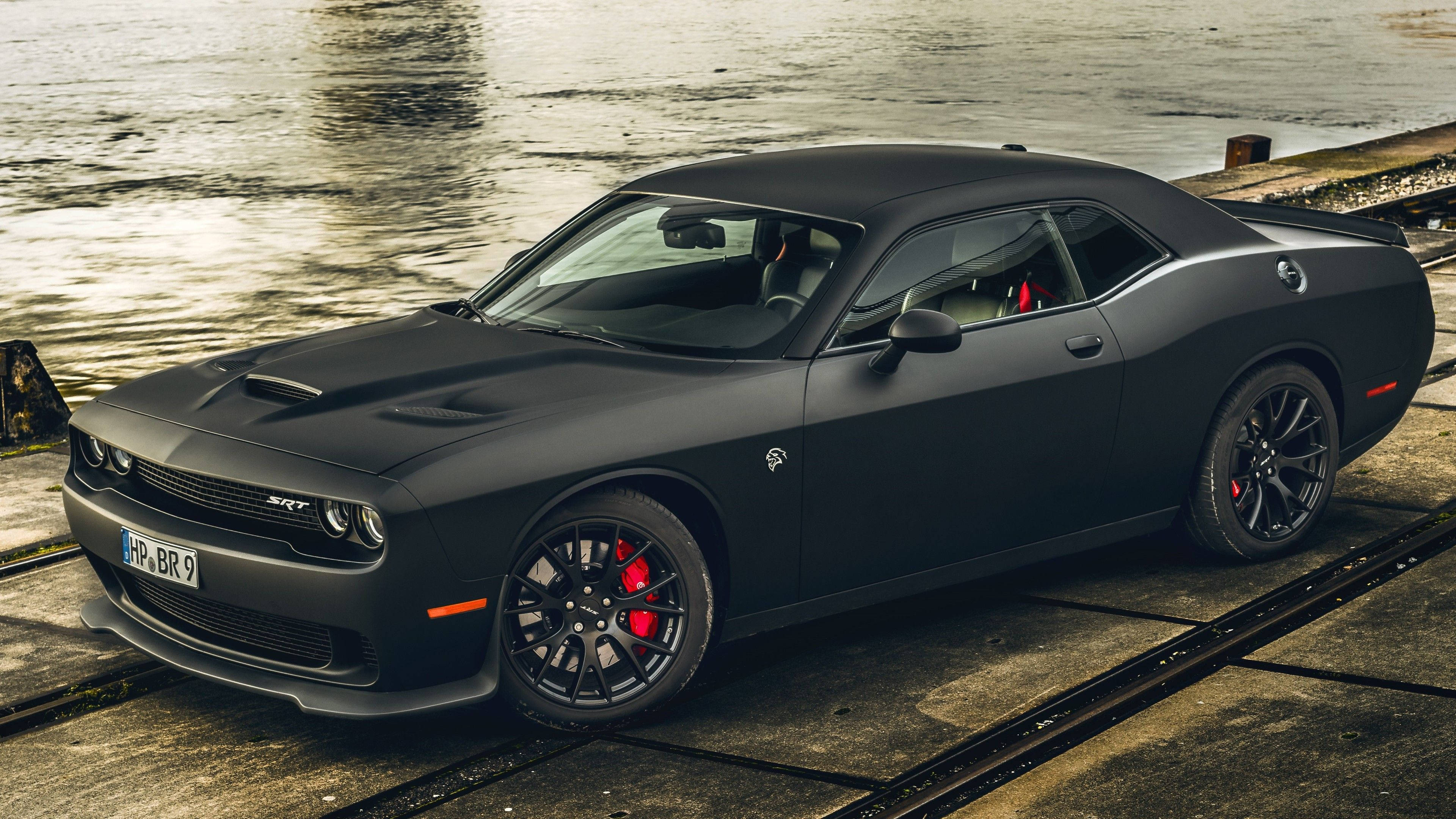 肌肉車 桌布 壁紙 — 一輛啞黑色Dodge Challenger SRT停放在近水的濕混凝土上，紅色煞車卡鉗清晰可見。