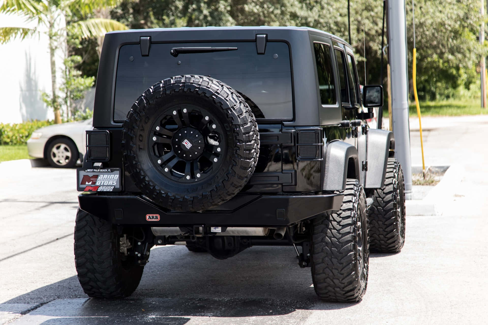 黑色吉普車 桌布 壁紙 — 一輛啞黑色升高的Jeep Wrangler，配備超大輪胎和車尾安裝的備用輪胎，停放在陽光明媚的瀝青停車場。