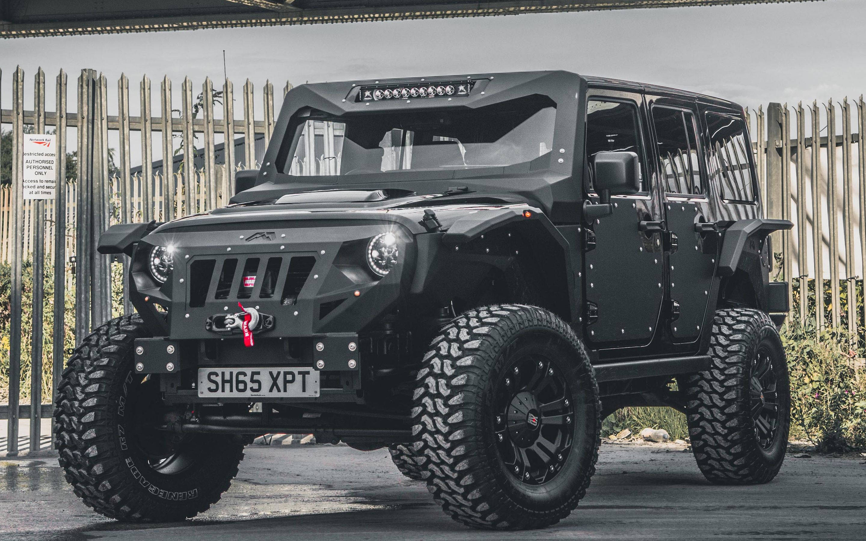 Wrangler 桌布 壁紙 — 一輛啞黑色堅固越野車配備特大輪胎，停泊在木柵欄附近，天空多雲。