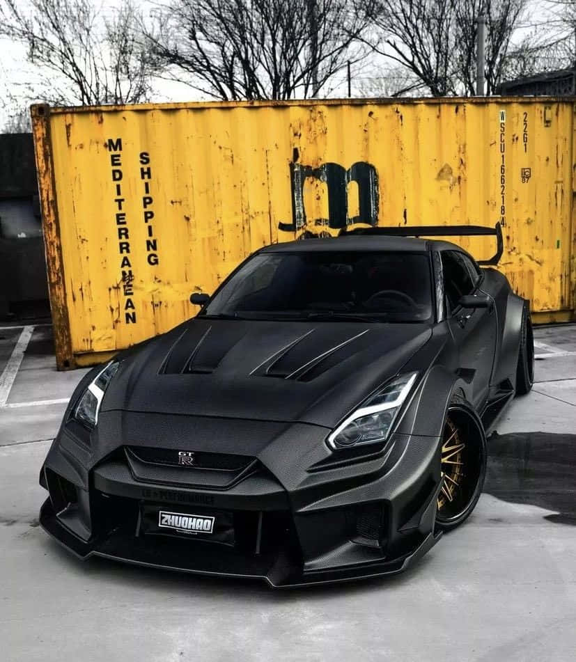Nissan 桌布 壁紙 — 一輛啞黑色改裝日產GT-R跑車，搭配金色輪圈，停放在一個風化的黃色貨櫃前面。
