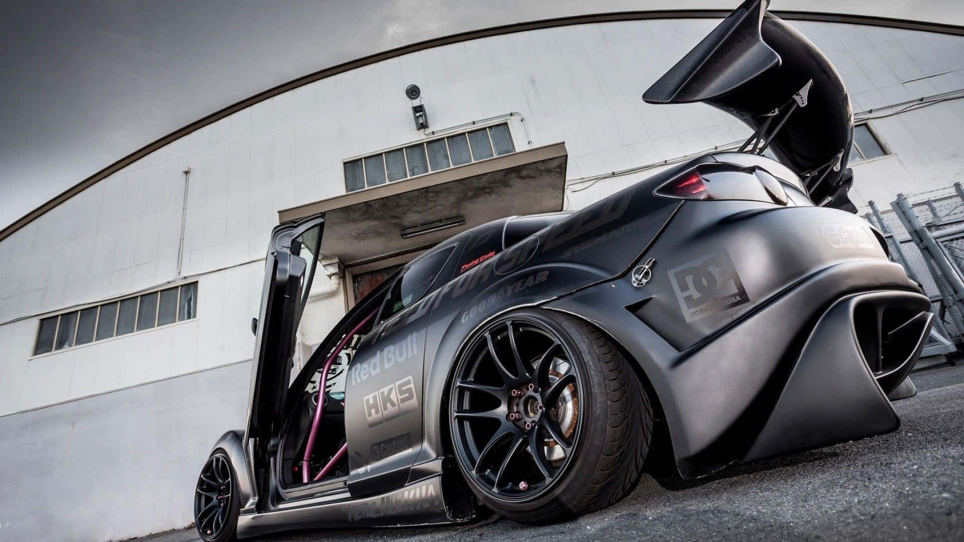 Mazda Rx-8 桌布 壁紙 — 一輛啞黑色改裝跑車，車門打開，帶有大型尾翼，停放在彎曲的工業建築前。