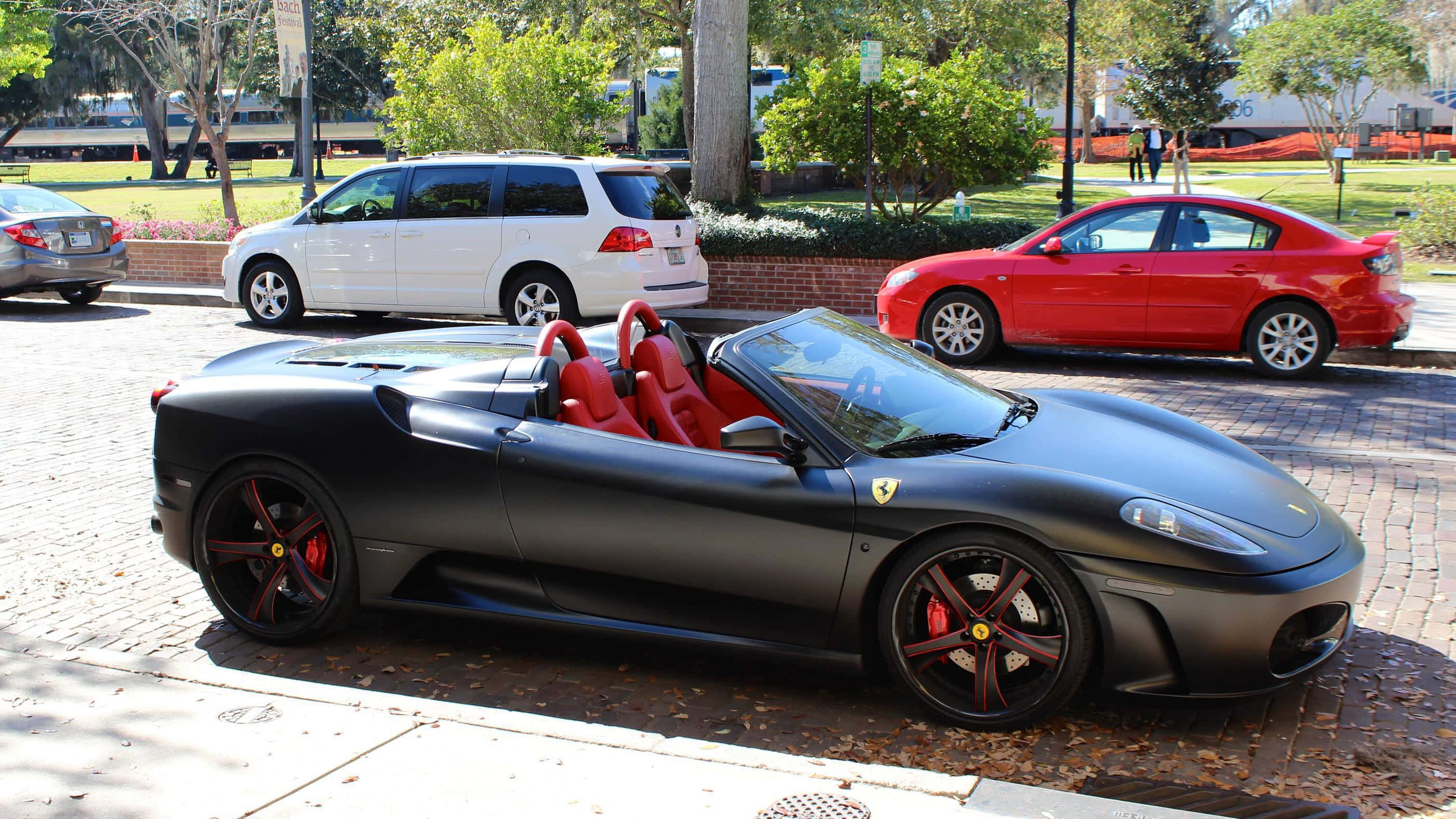 Ferrari F430 桌布 壁紙 — 一輛啞黑色法拉利Spider，配備紅色內裝和煞車卡鉗，在明亮的日光下停放在磚街旁的其他車輛旁邊。