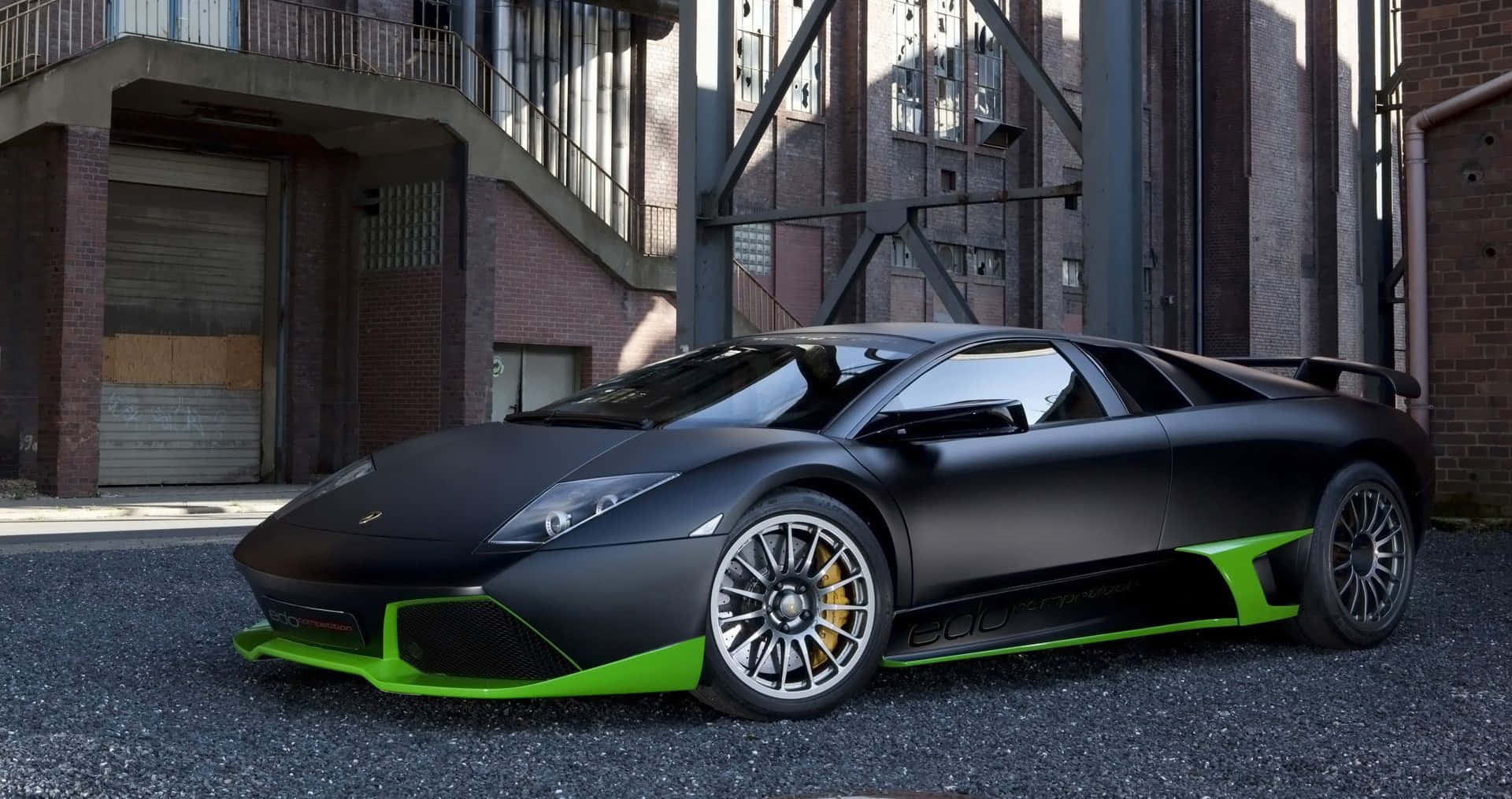 Lamborghini Murciélago 桌布 壁紙 — 一輛啞黑色藍寶堅尼搭配螢光綠色飾邊，停在工業磚造建築前的碎石地上。