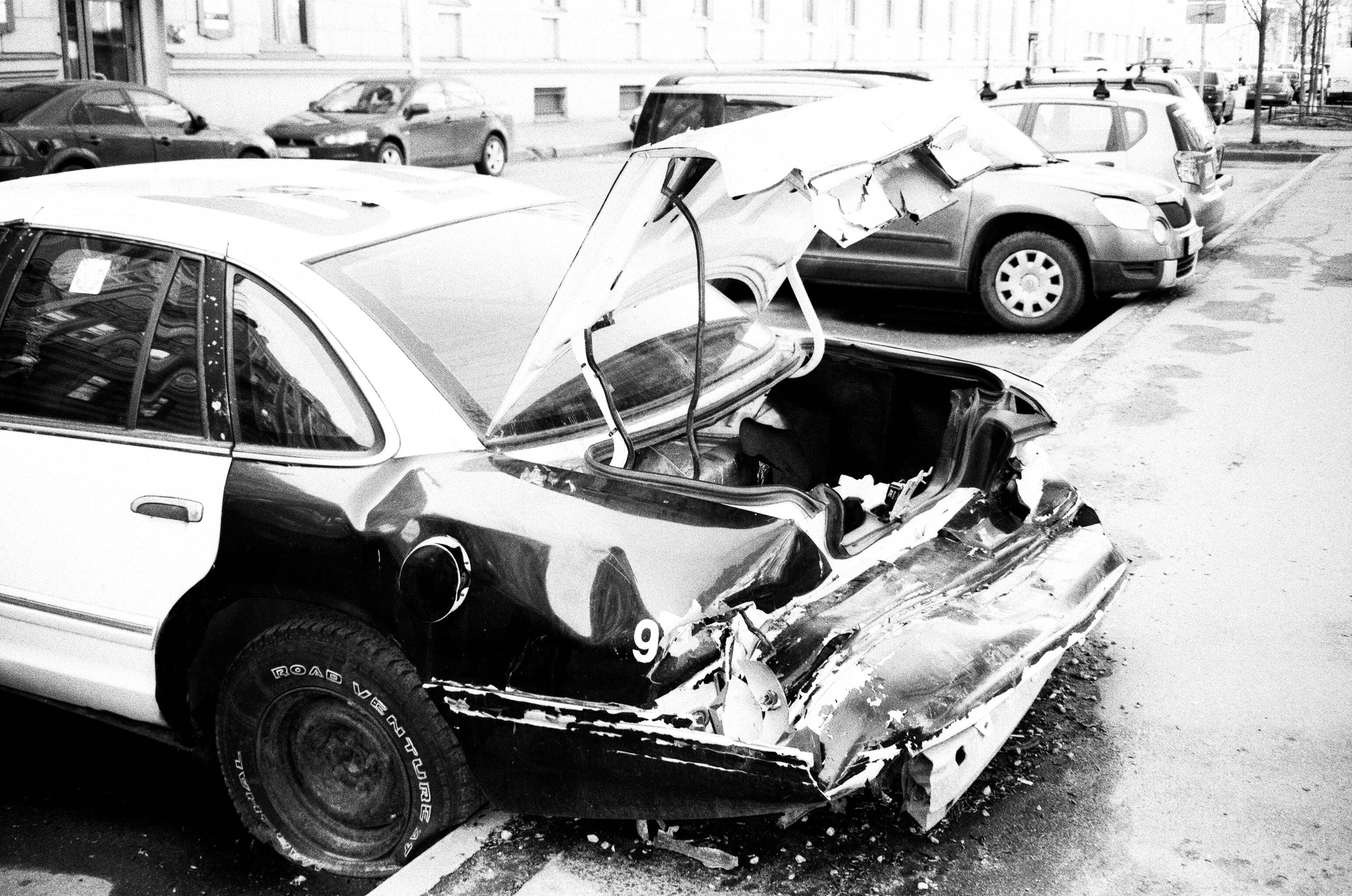 黑白 桌布 壁紙 — 一輛嚴重損壞的汽車，車尾被壓碎，後車廂打開，被遺棄在潮濕的城市街道上，黑白照片。