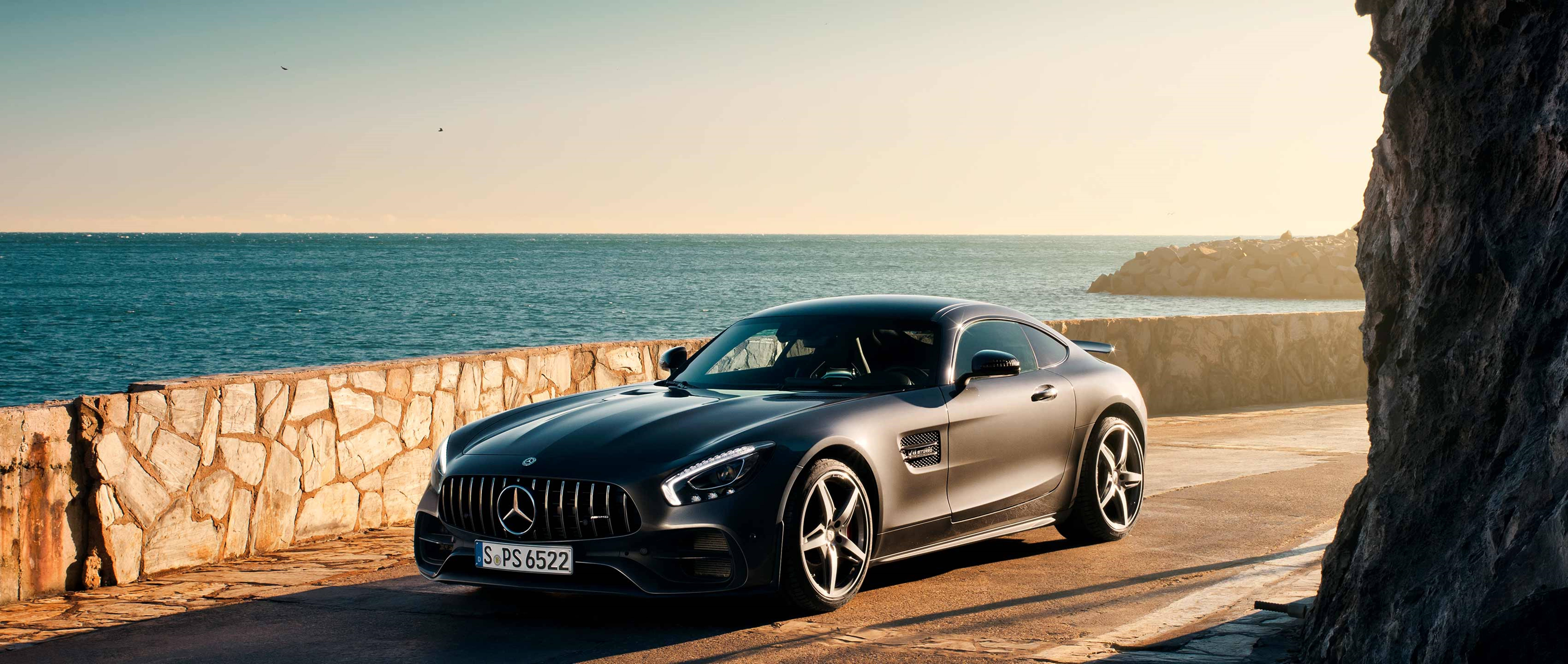 Mercedes 桌布 壁紙 — 一輛型格啞光灰色賓士AMG GT跑車停泊在陽光普照的海岸道路旁，旁邊有石牆和海洋。