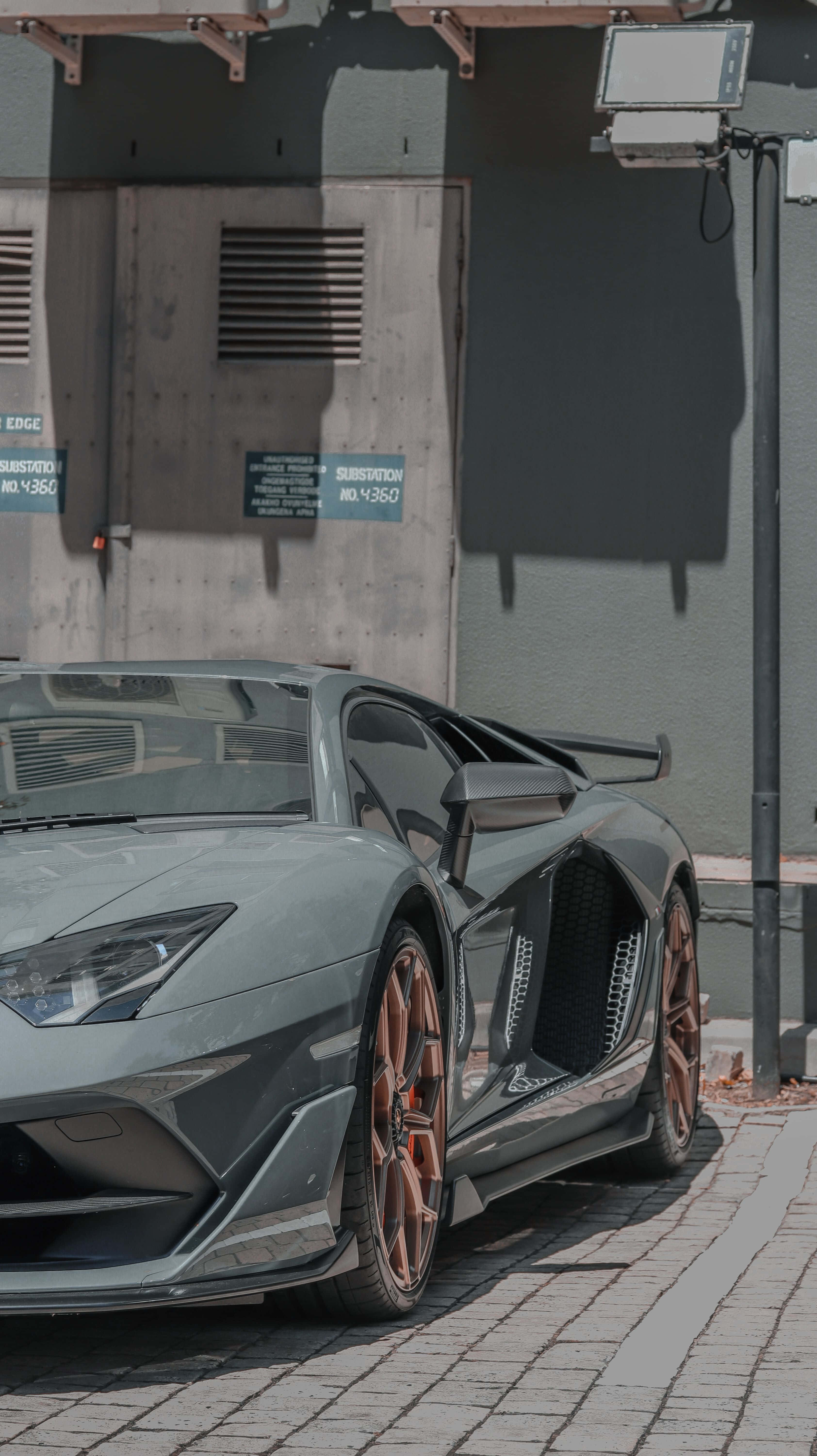 Minimalist Car 桌布 壁紙 — 一輛型格消光灰色藍寶堅尼Aventador搭配銅色輪圈，停放在陽光普照的磚砌人行道上，旁邊是混凝土建築物。