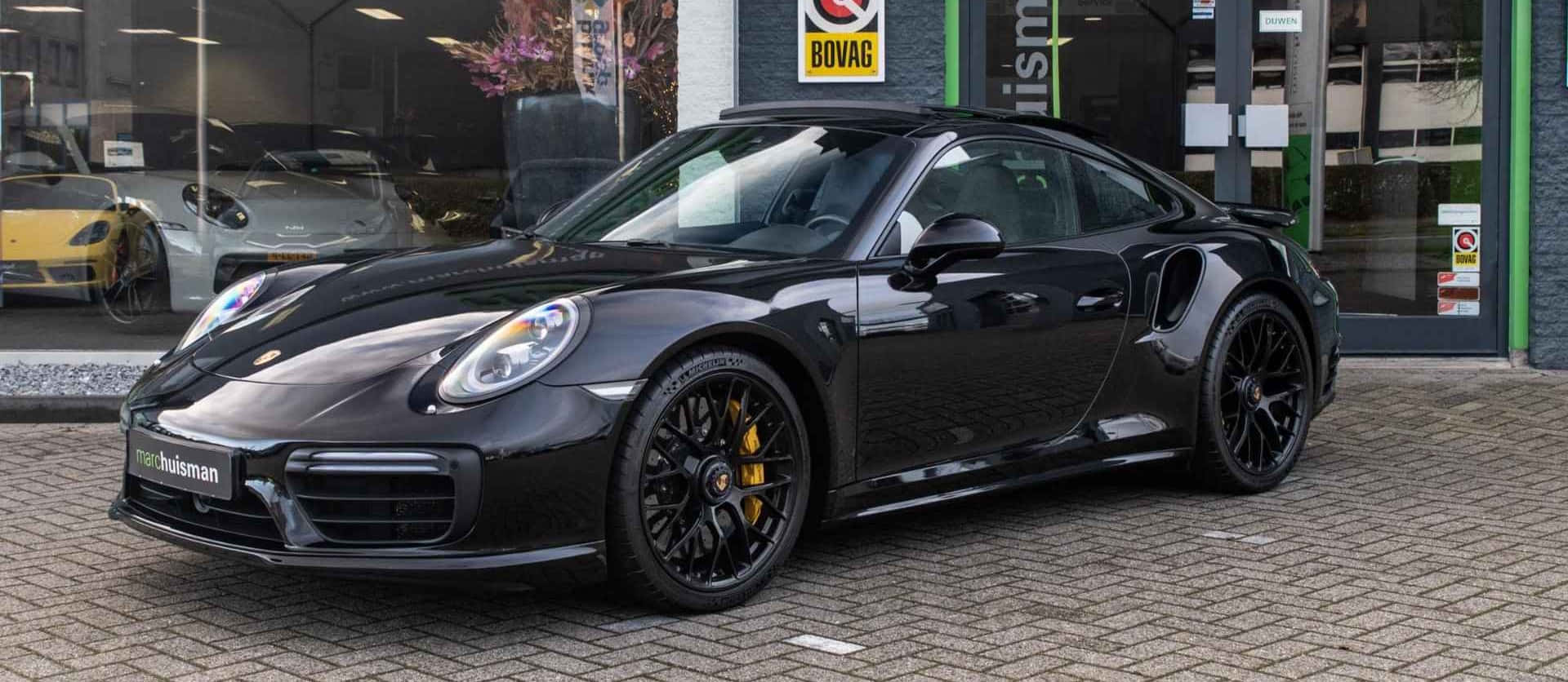 Black Porsche 911  – 4K Car Wallpaper 壁紙 — 一輛型格黑色保時捷911 Turbo S，配備黃色制動卡鉗，停在汽車經銷商前的磚砌車道上。