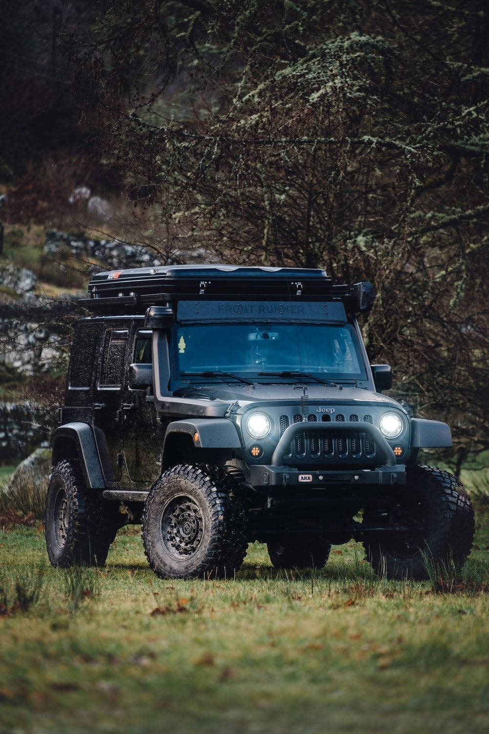 Black Jeep 桌布 壁紙 — 一輛堅固的啞光灰色Jeep Wrangler，配備車頂行李架和明亮頭燈，停泊在長滿草的地形上，周圍是光禿禿的樹木。