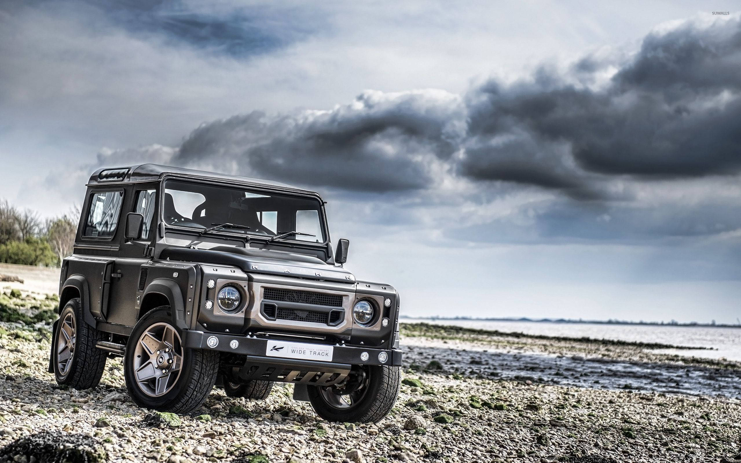 荒原路華 桌布 壁紙 — 一輛堅固的啞光黑色 Land Rover Defender 停在岩石海岸線上，天空多雲，氣氛戲劇化。