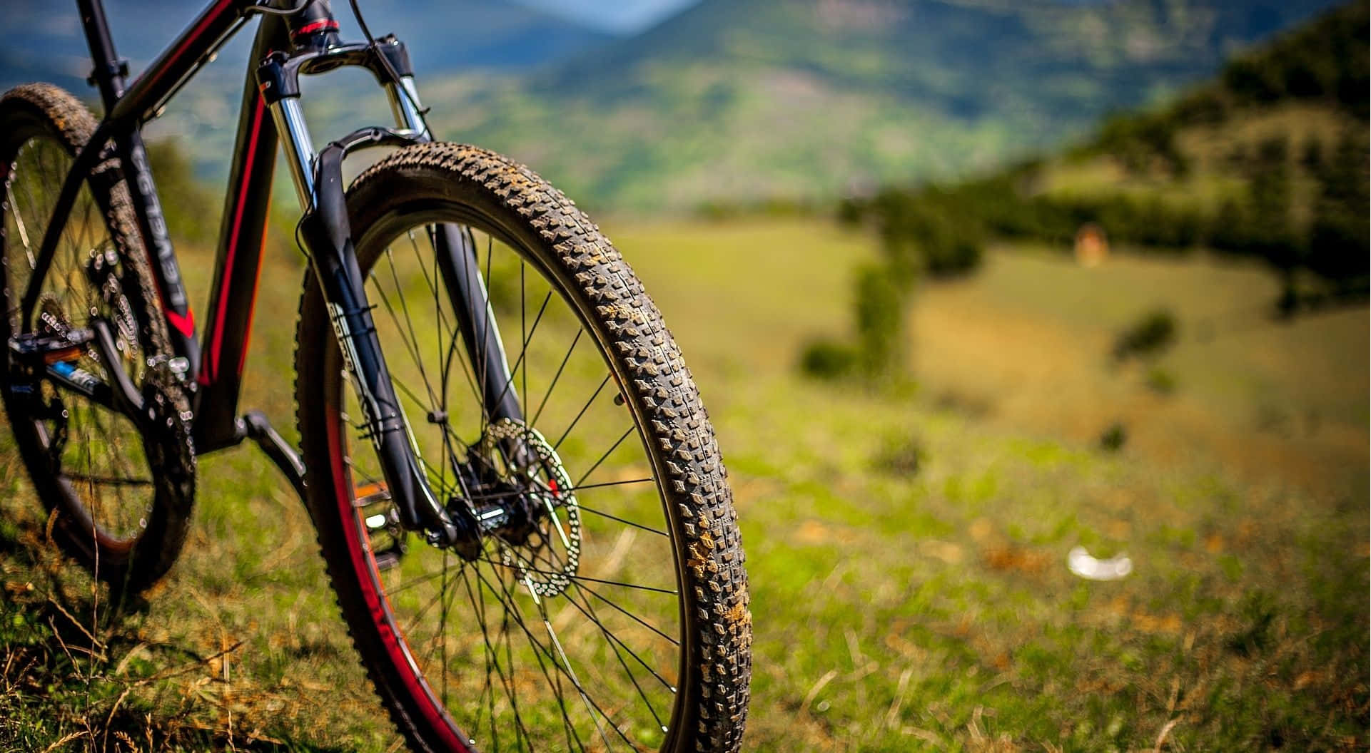 Mtb 桌布 壁紙 — 一輛堅固的山地自行車，輪胎沾滿泥濘，停在草地地形上，背景是起伏的綠色山丘和遠處的山脈。