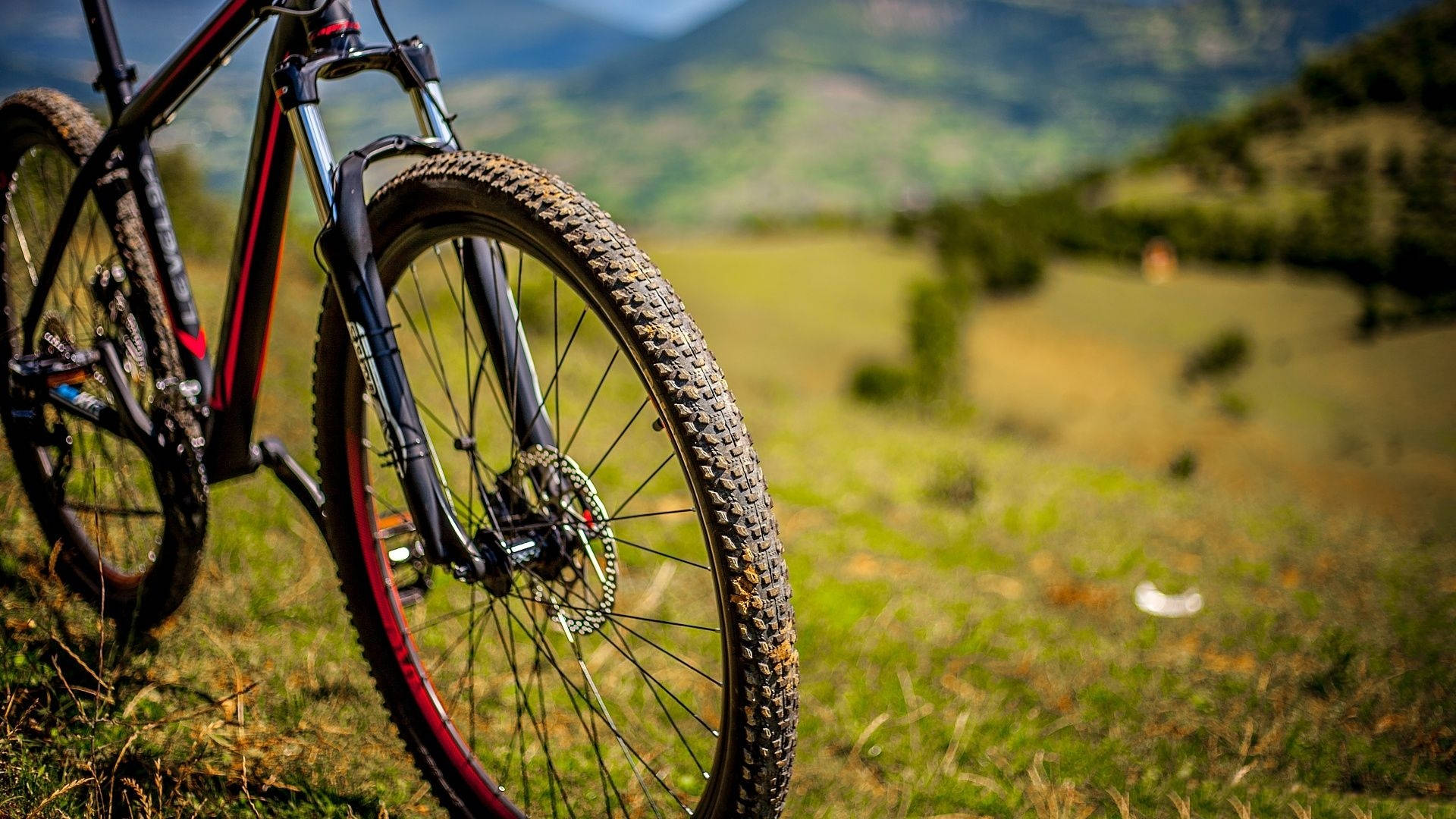 Mountain Bike 桌布 壁紙 — 一輛堅固的山地車，輪胎沾滿泥濘，停放在草地上，背景是起伏的綠色山丘和遠山。