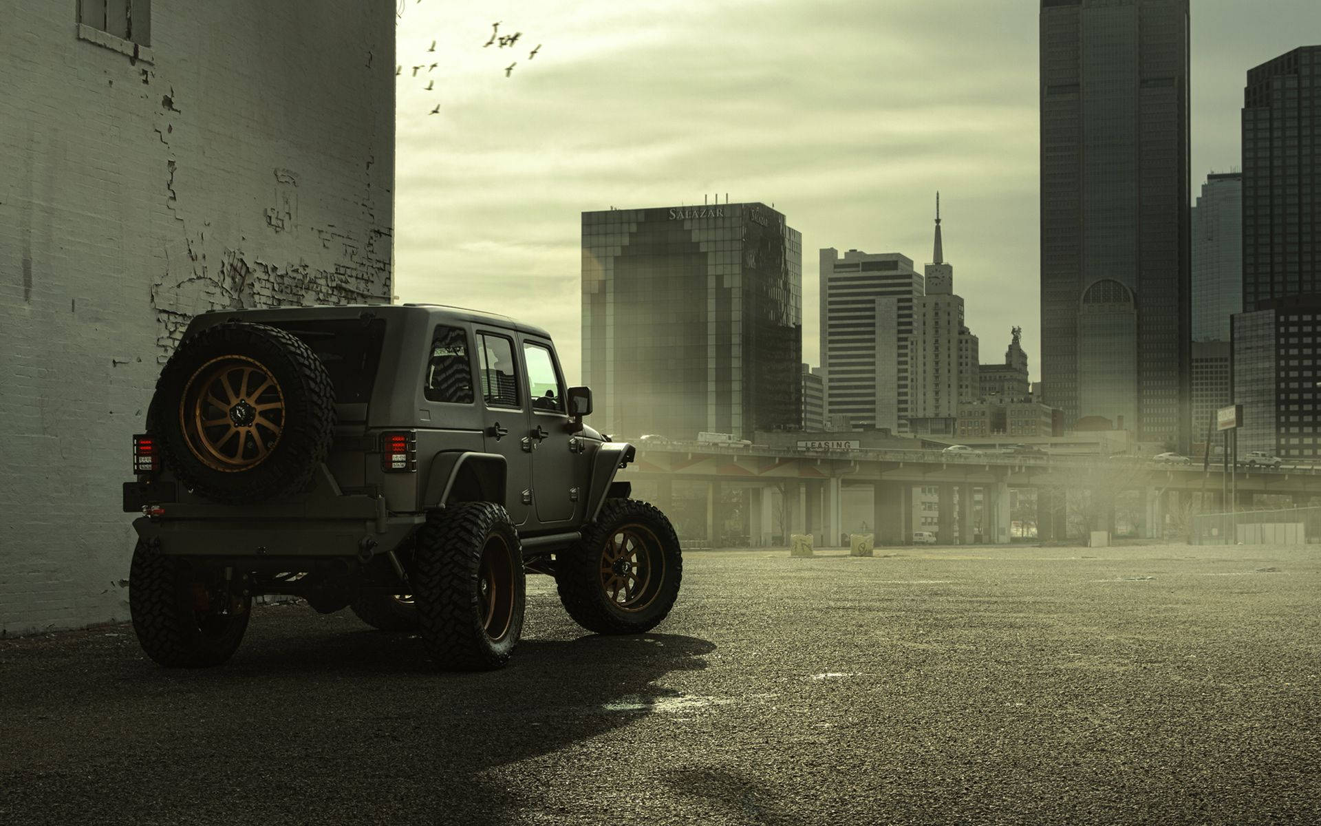 Wrangler 桌布 壁紙 — 一輛堅固的橄欖綠色Jeep Wrangler配金色輪圈，停放在空曠的城市空地，背景是煙霧瀰漫的陰天下高聳的摩天大樓。