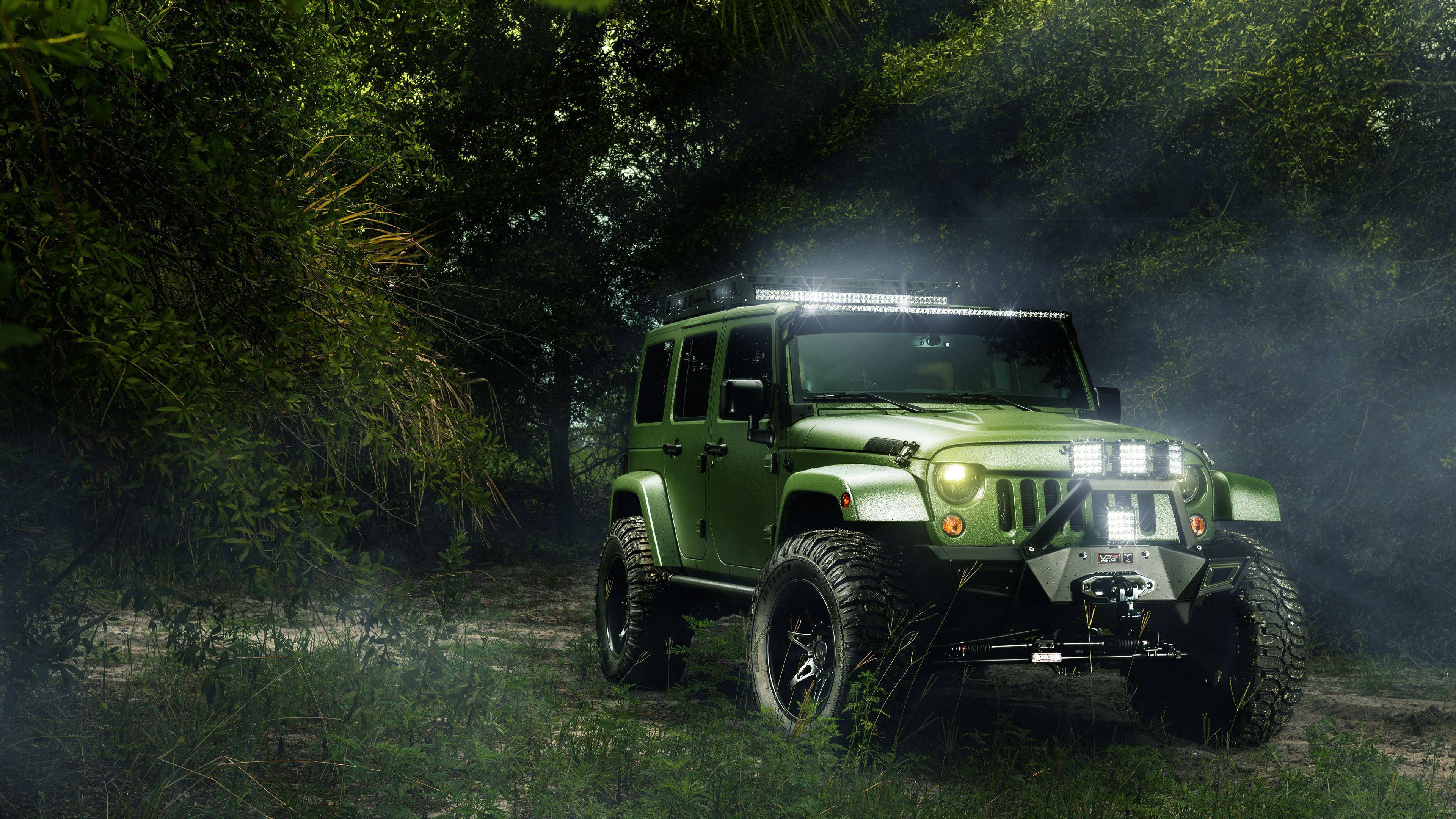 汽車 桌布 壁紙 — 一輛堅固的橄欖綠色Jeep Wrangler在夜間穿過霧氣瀰漫的森林，明亮的車頭燈照亮四周，周圍是茂密的樹葉。